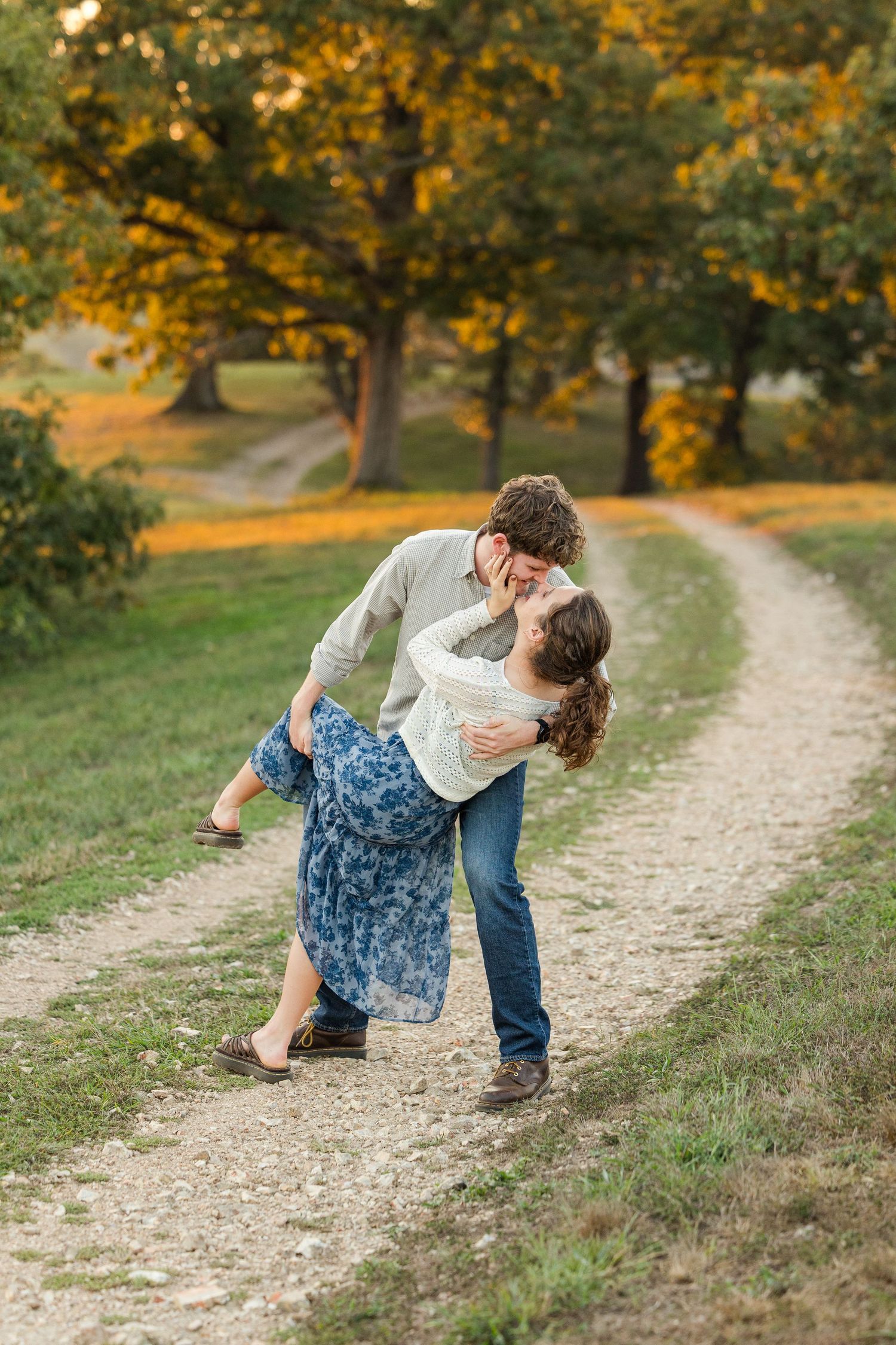 A romantic couple dances and dips along a winding dirt path surrounded by golden autumn trees at sunset.