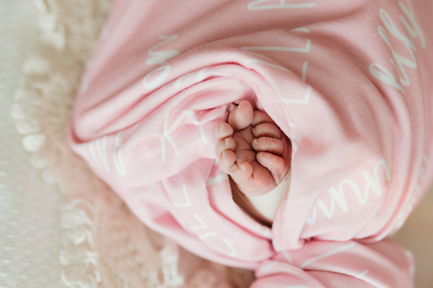 A tiny baby foot peeks out from soft pink swaddling blanket against a white lace background.