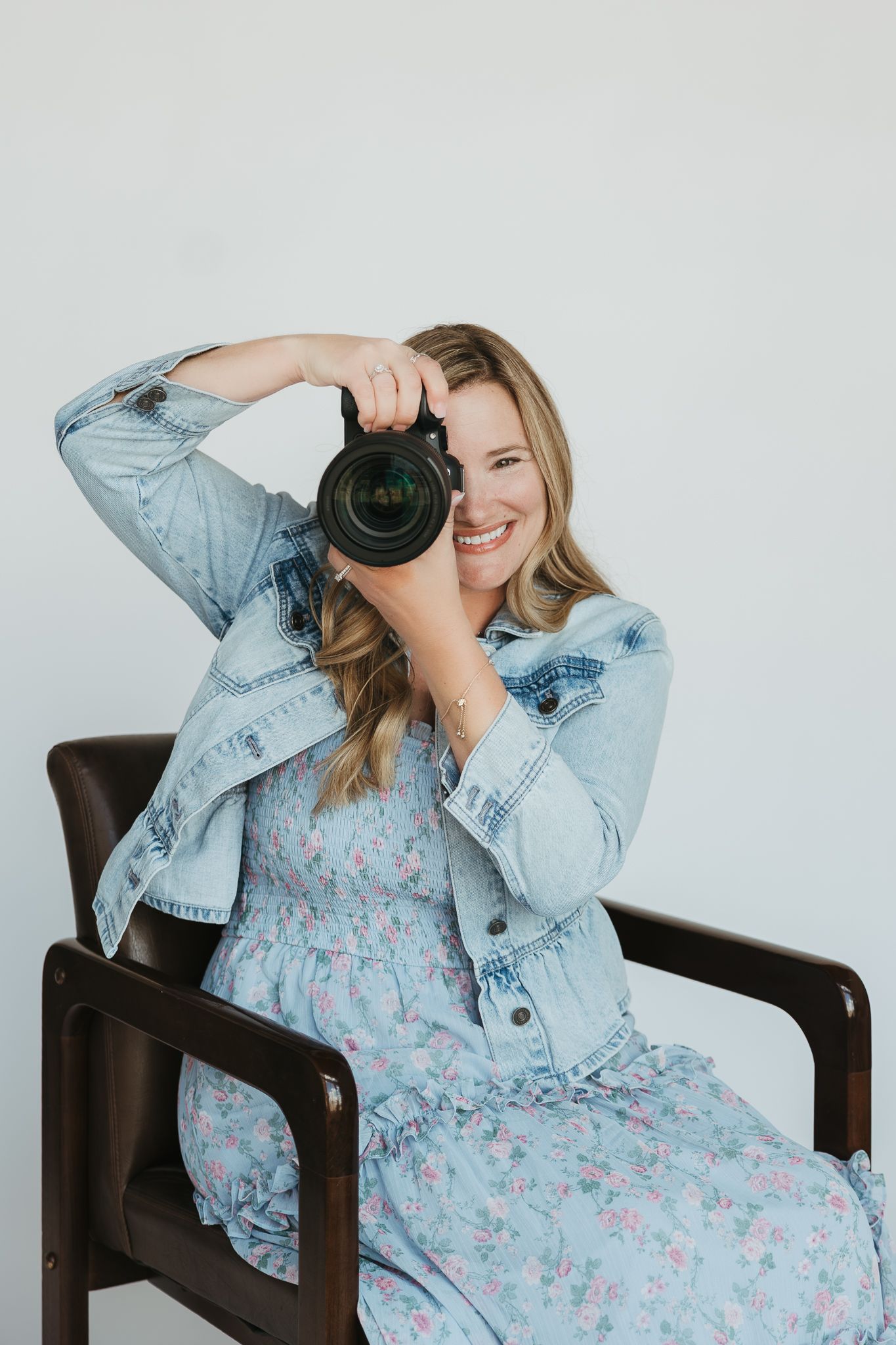 Person in floral dress and denim jacket poses with camera in chair during lifestyle photoshoot.