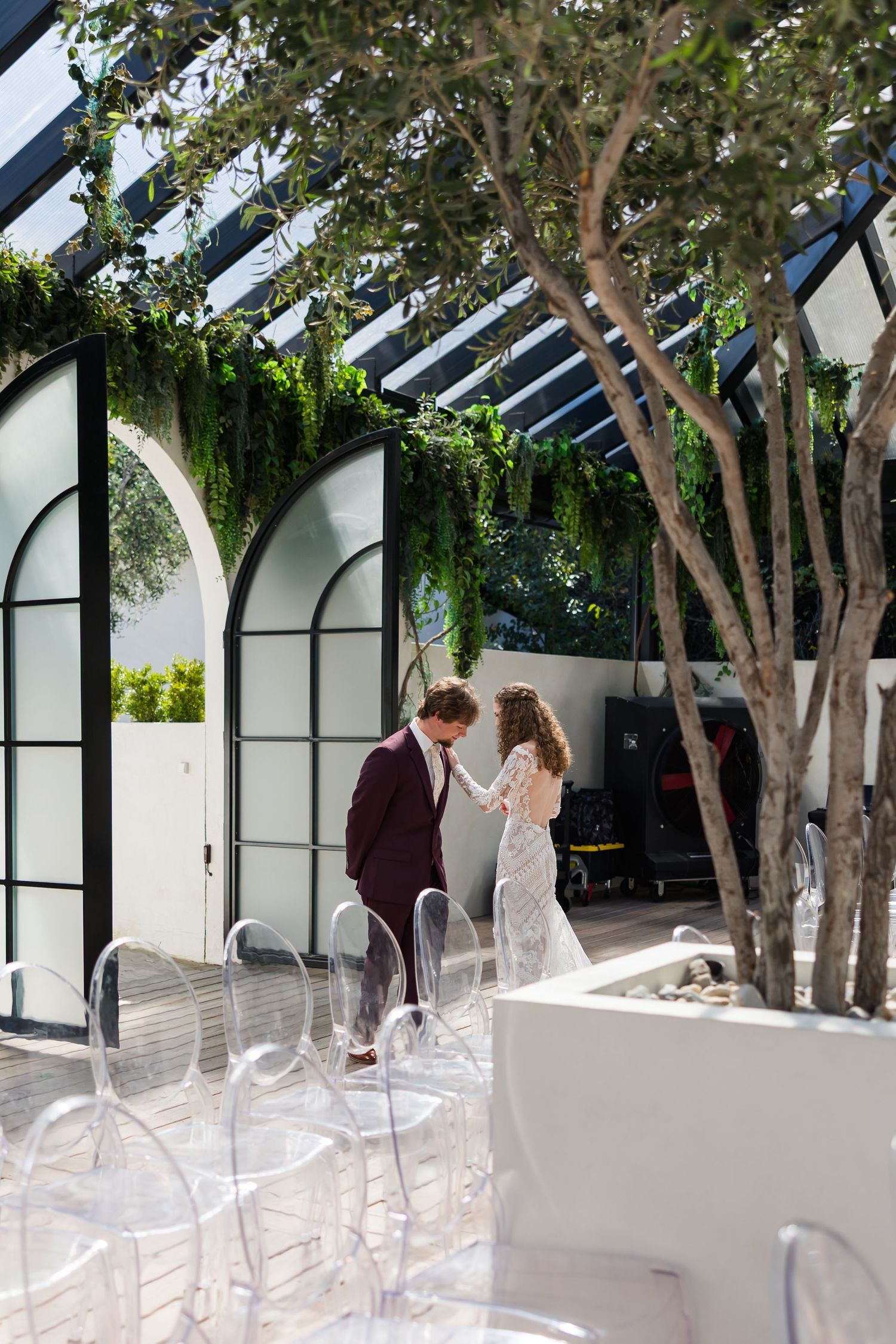 Bride and groom share a tender moment holding hands in a sunlit greenhouse venue with arched windows and lush greenery.