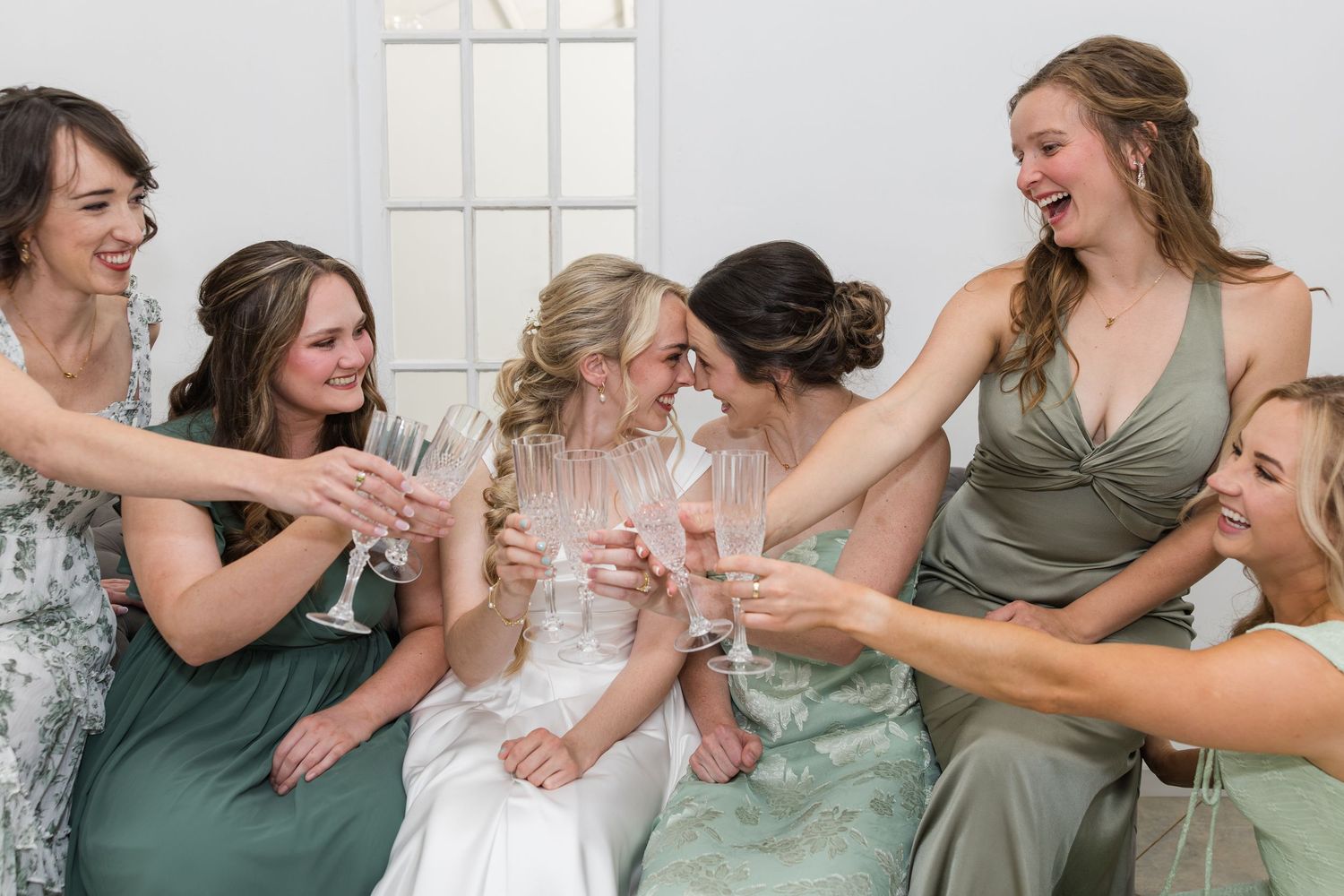 Bride and bridesmaids in sage green dresses laughing and toasting with champagne glasses at a wedding celebration.
