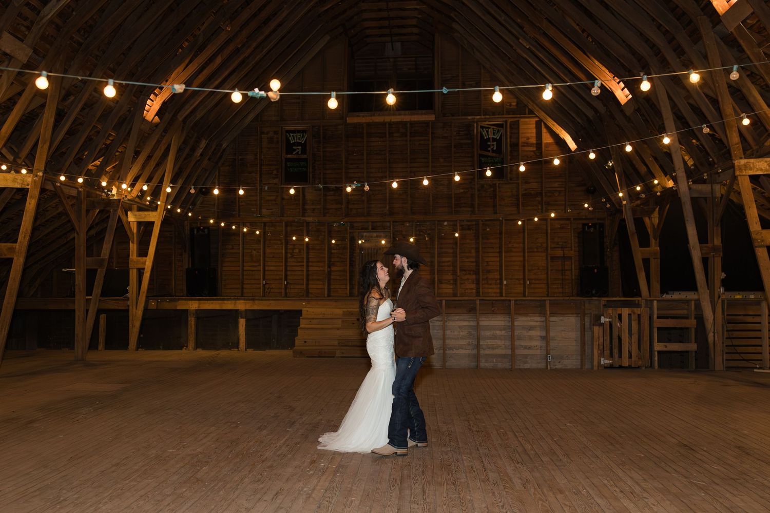 Bride and groom sharing first dance in a warmly lit rustic barn adorned with string lights at wedding reception.