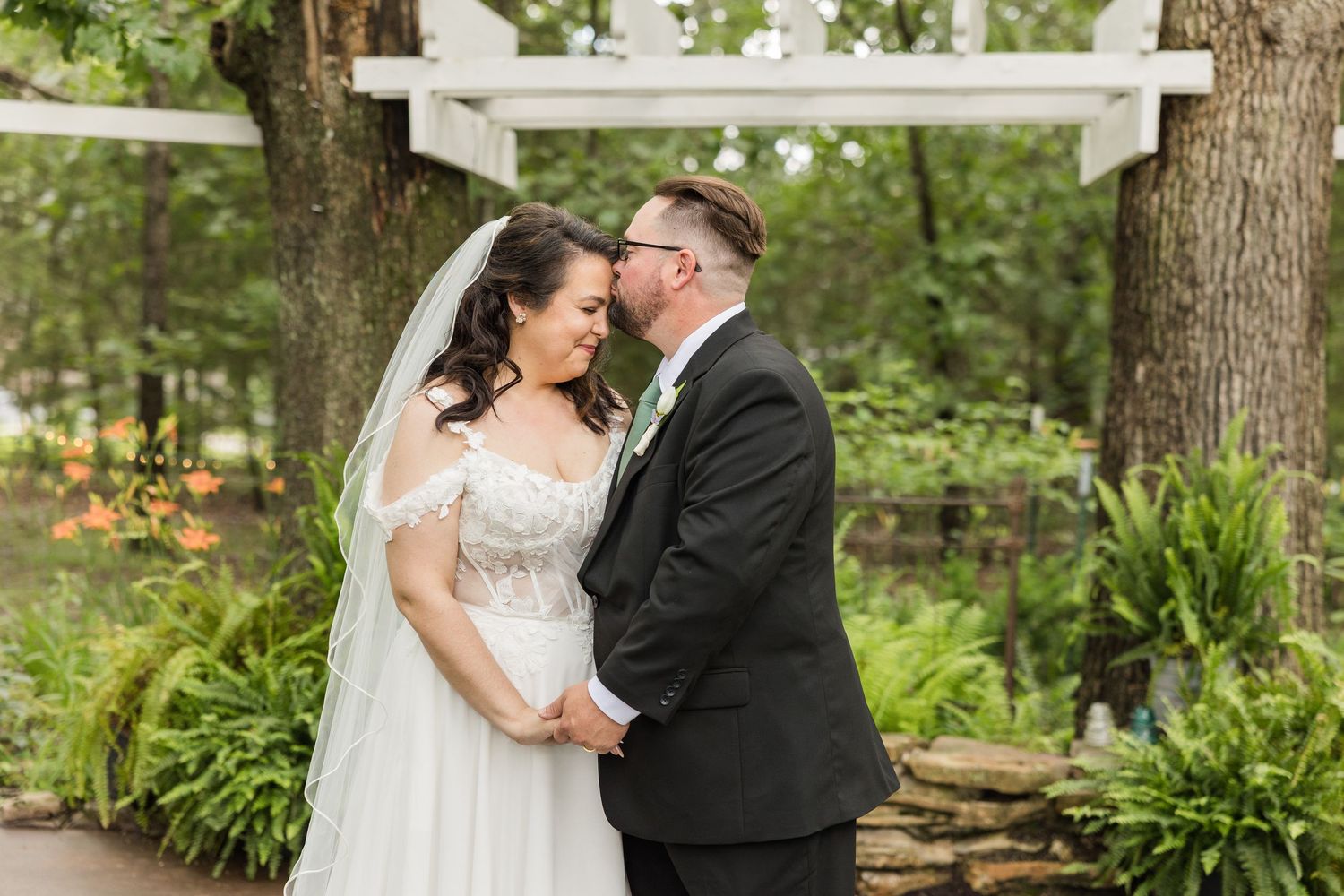 Bride and groom share a romantic moment under a white arbor surrounded by lush greenery at an outdoor garden wedding.
