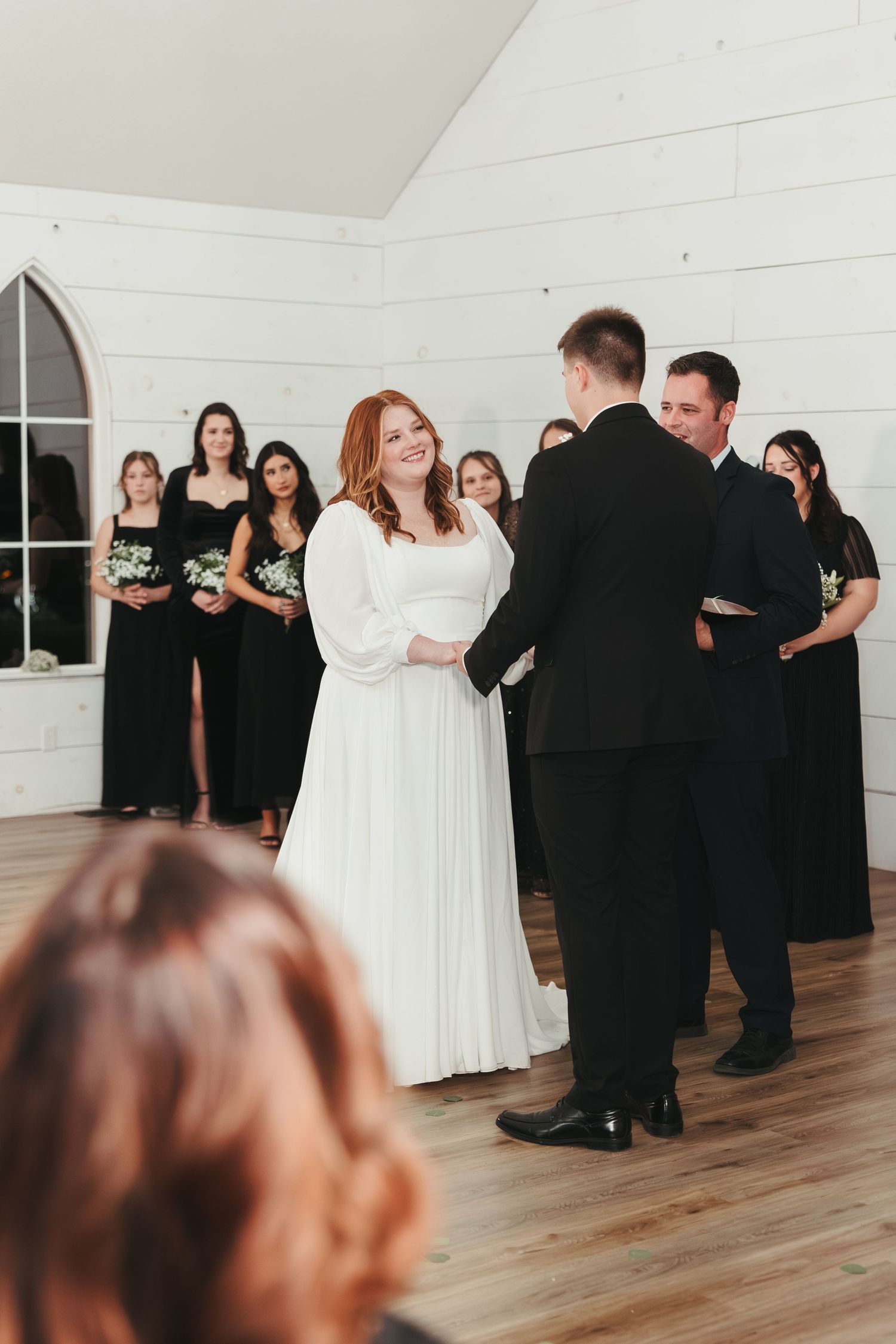 A wedding ceremony with bridesmaids in black dresses and a bride in a white gown standing at the altar.