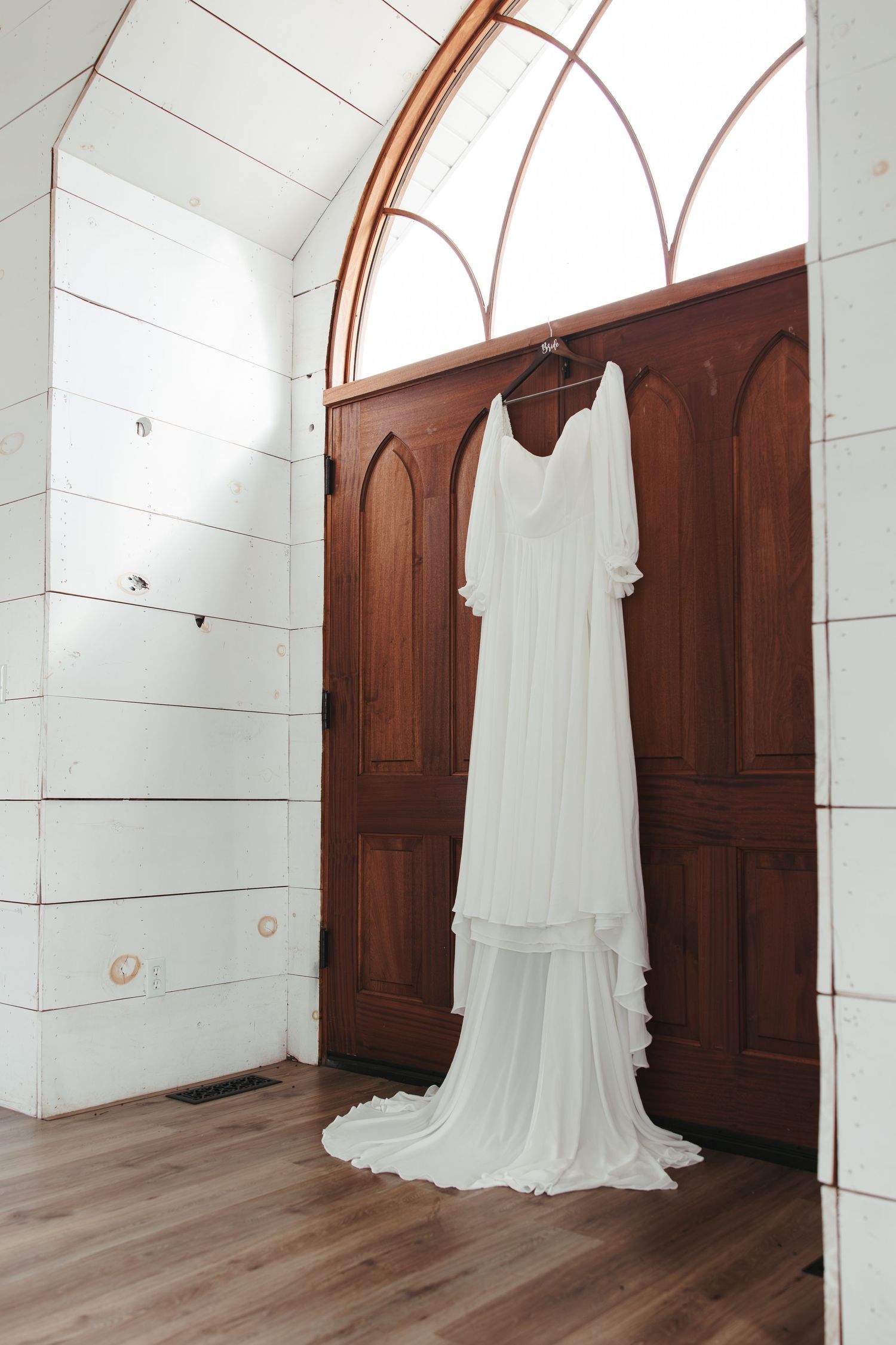 Elegant white wedding dress hanging in church doorway with arched wooden details.
