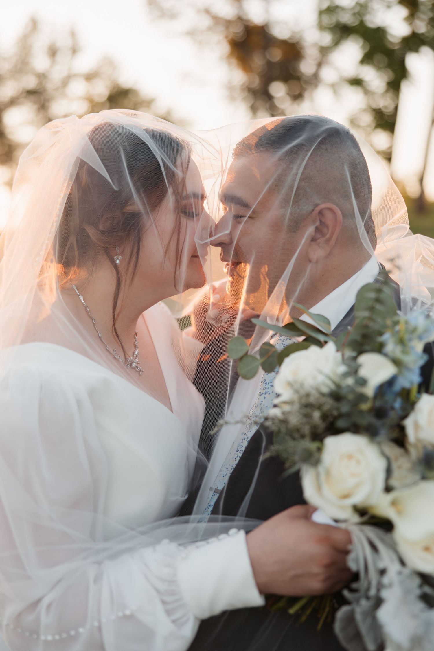 A romantic wedding moment under a delicate veil with a white rose bouquet in golden sunlight.