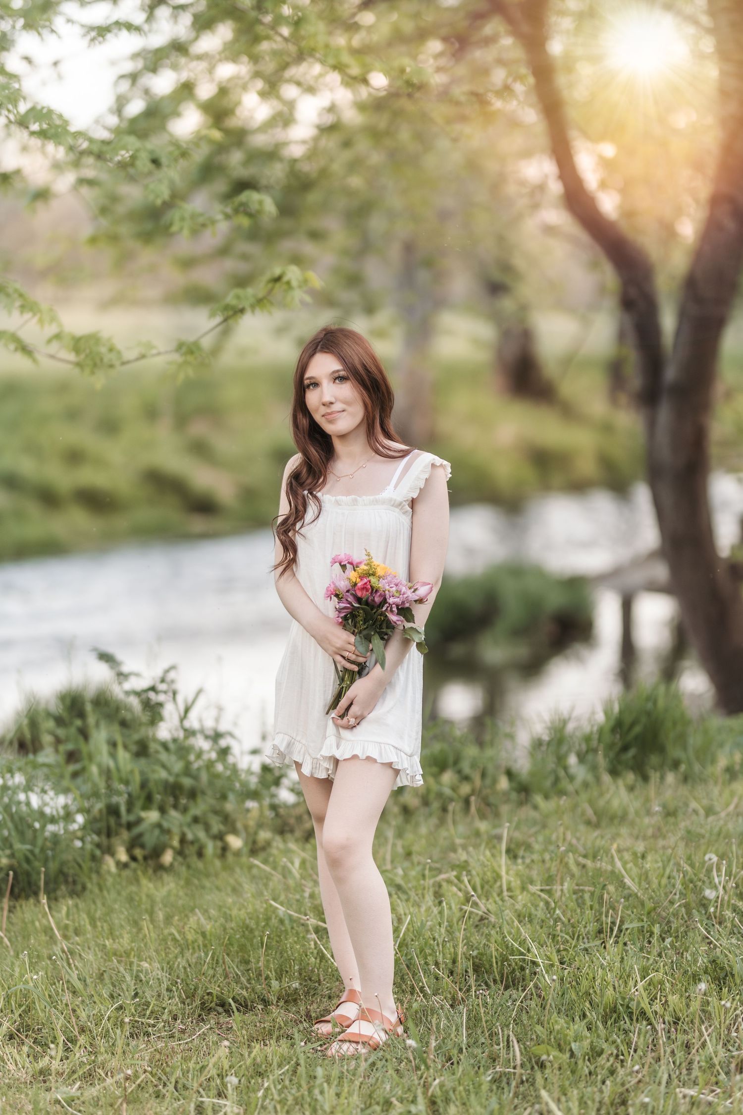 A person in a white dress holds flowers while standing in a sunlit meadow near a river.