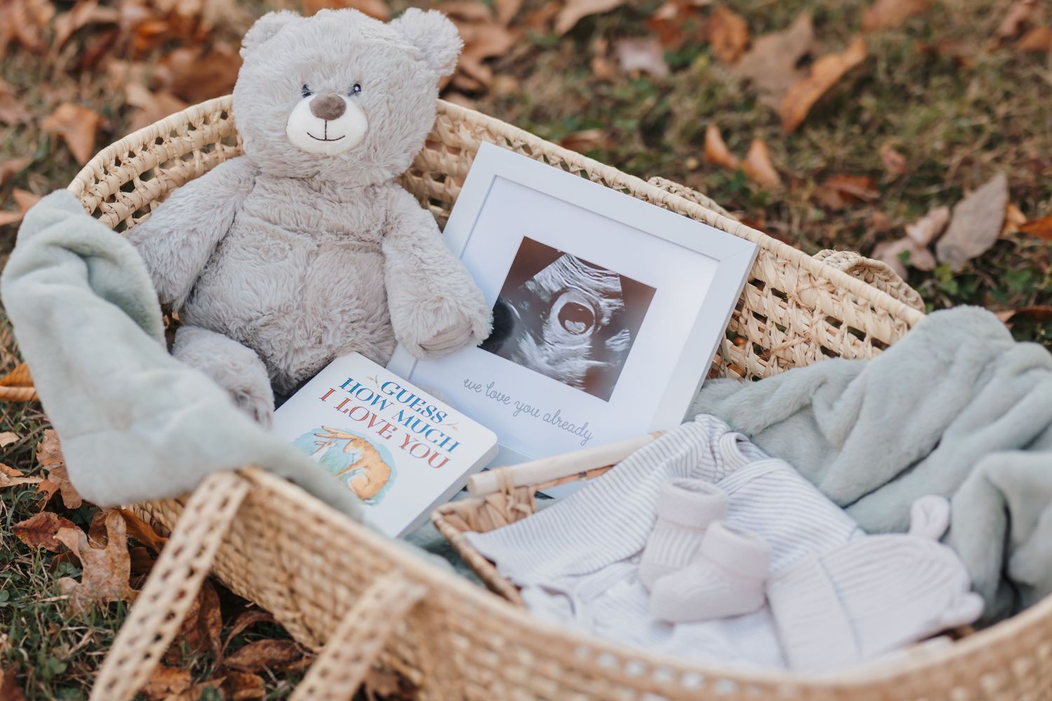 Baby announcement setup with teddy bear, ultrasound photo, and soft items in wicker basket.