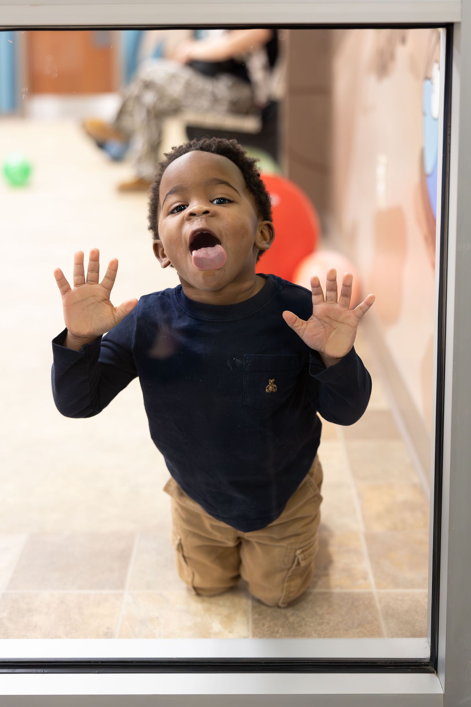 Young child in navy sweater showing excitement with raised hands and animated expression through a window.