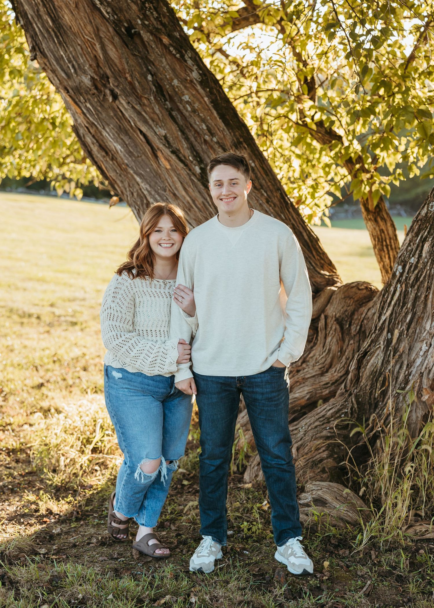 A couple poses together in front of a massive tree trunk in warm autumn lighting.