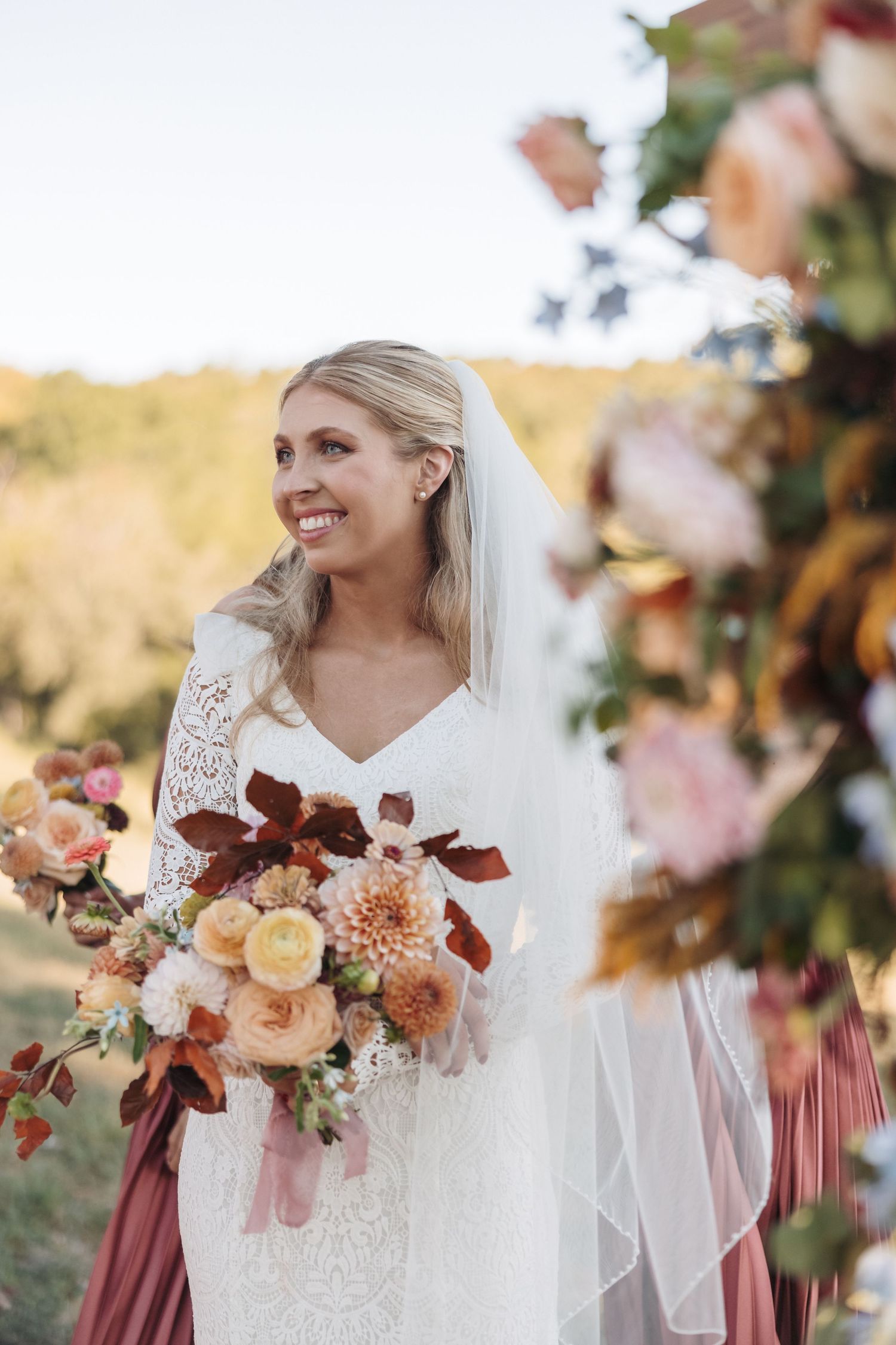 Elegant bride in lace dress holding autumn-colored bouquet with peach and burgundy blooms.