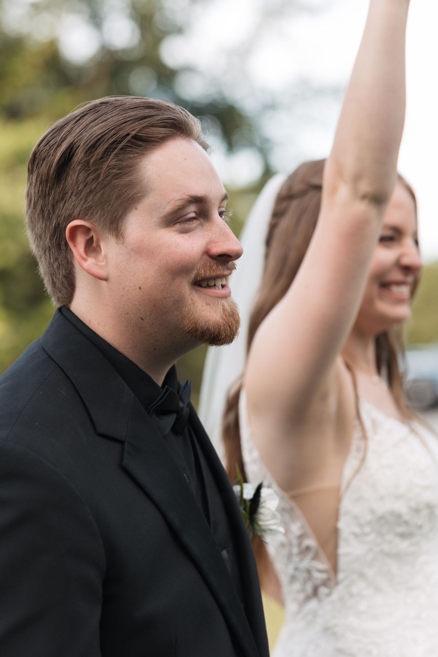 A couple at their wedding celebration expressing joy with raised arms outdoors.