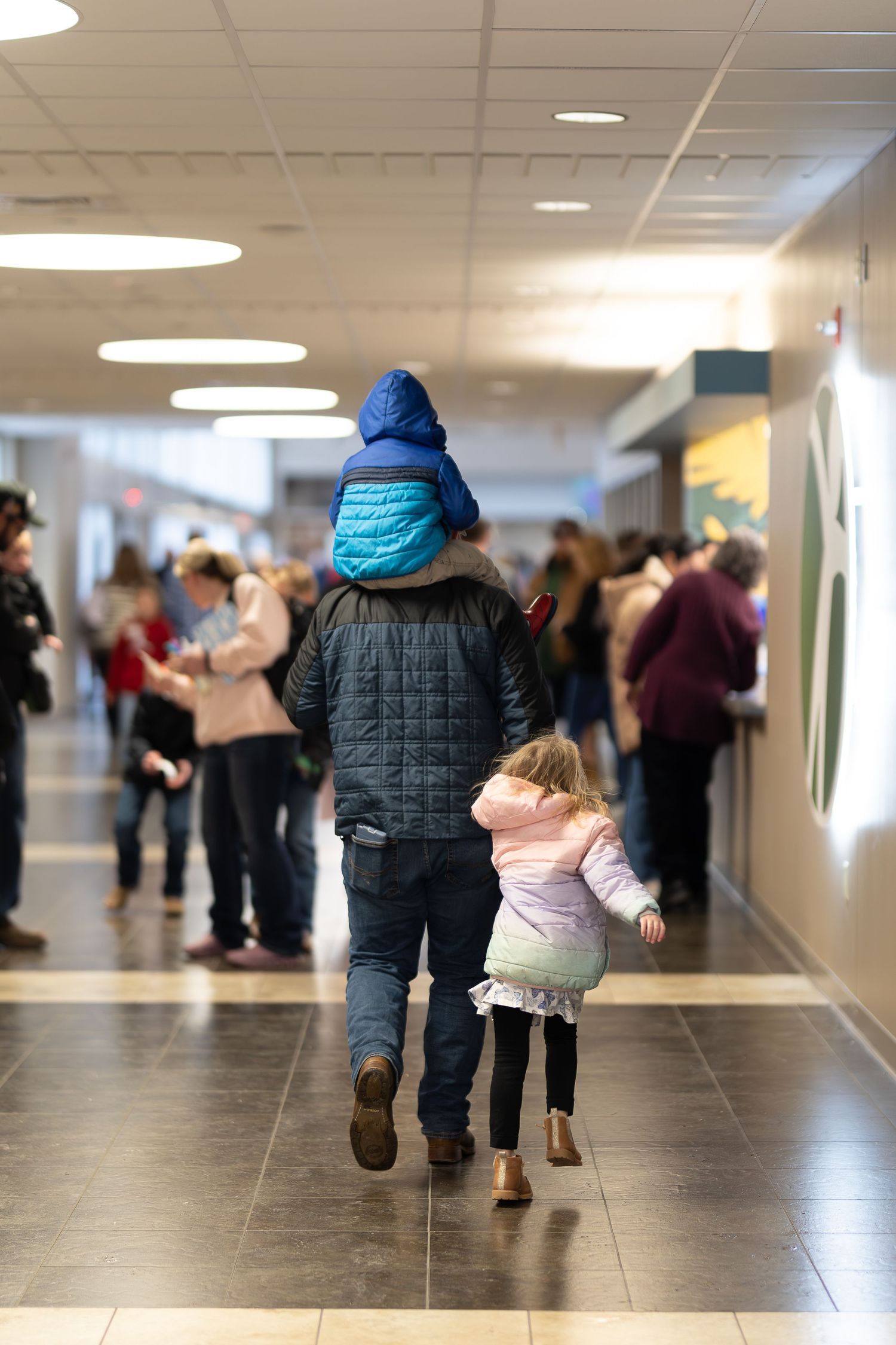 Two people walking down a busy indoor hallway with fluorescent lighting overhead.