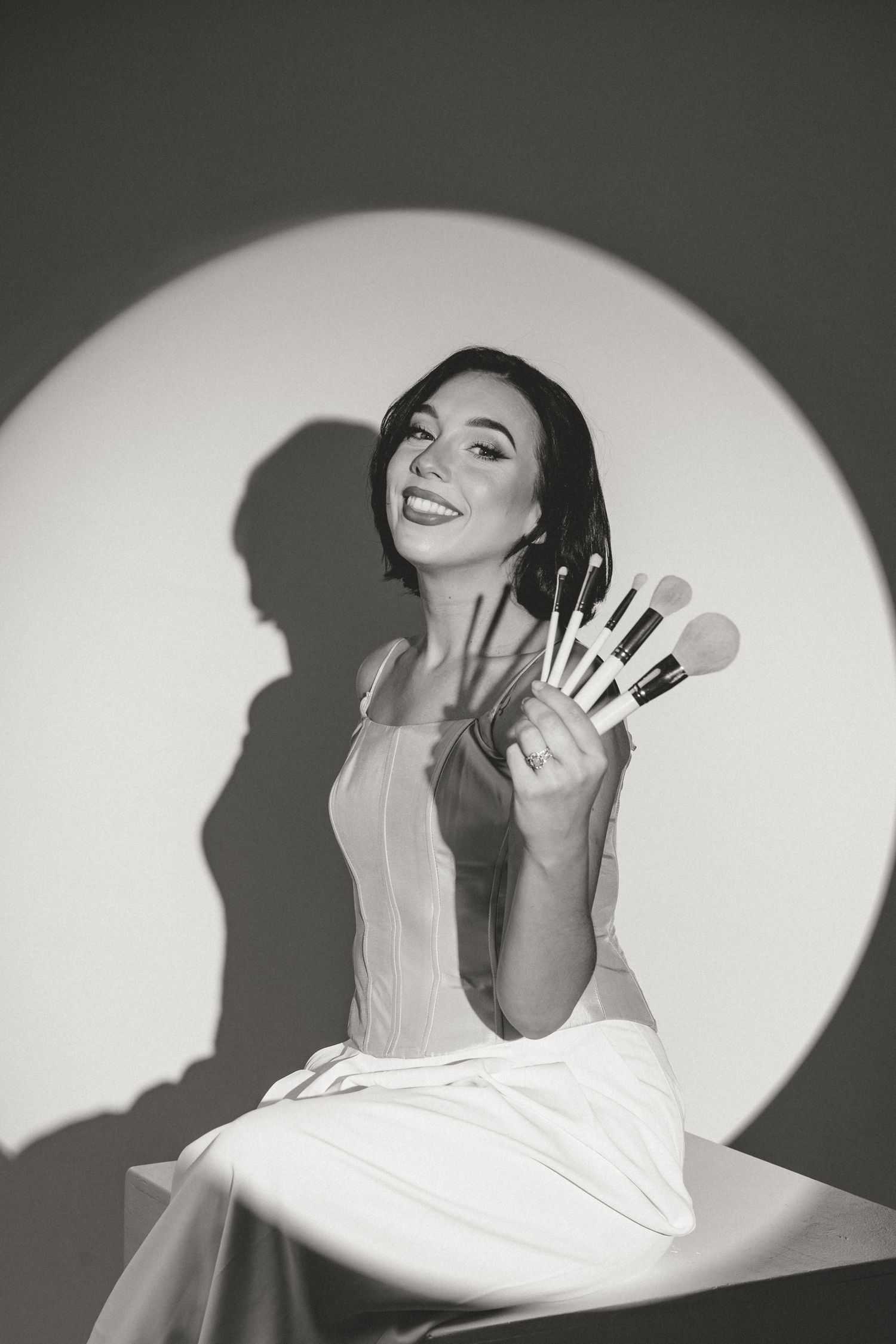 Black and white portrait holding makeup brushes against a spotlight backdrop.