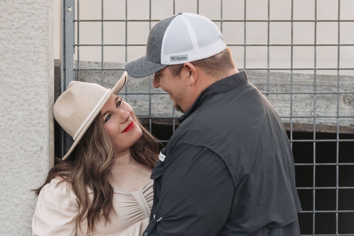 Intimate moment between couple with woman in beige hat and man in dark jacket against industrial backdrop.