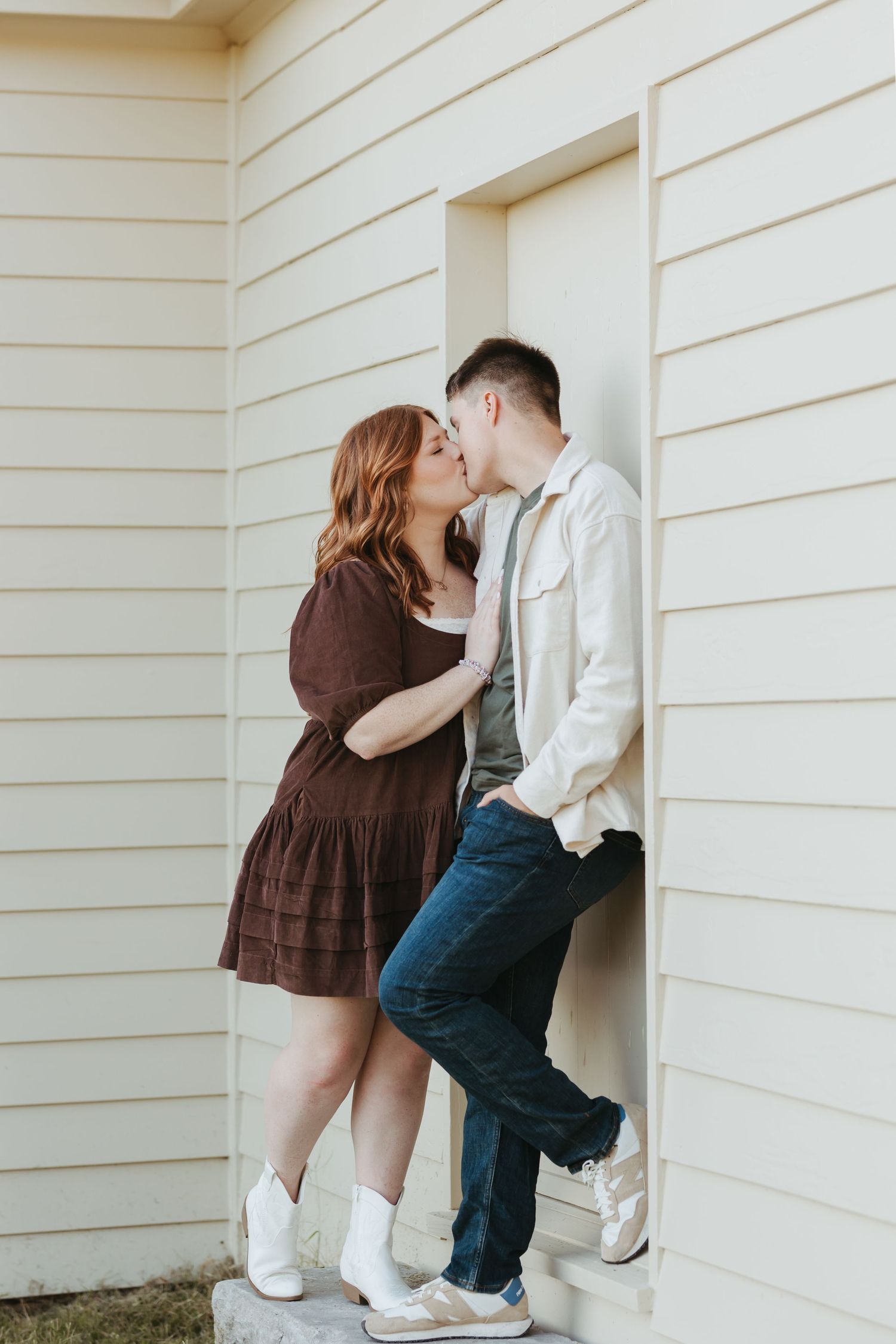 Intimate moment between couple against white siding wall, with woman in brown dress and man in light jacket.