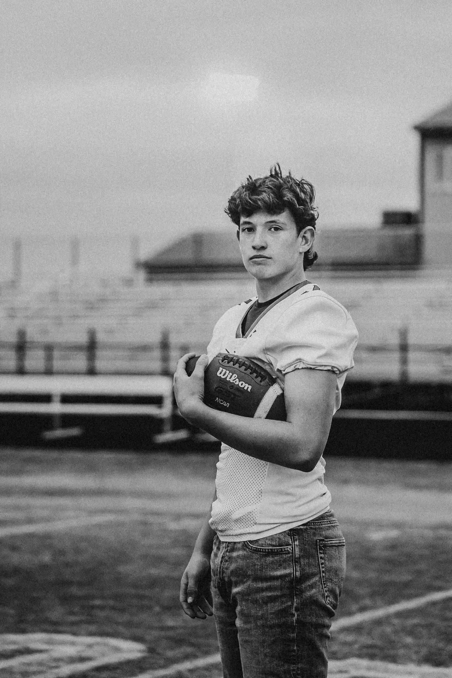 Black and white portrait of athlete holding football on empty field with stadium background.