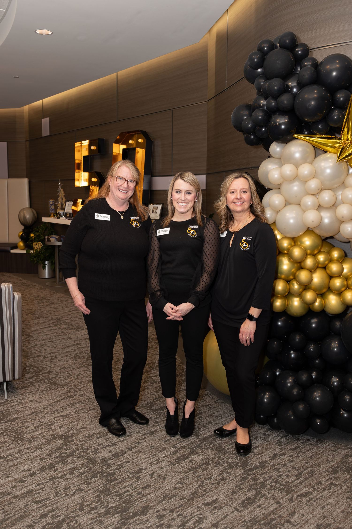 Staff members pose beside black and gold balloon decoration at corporate event.