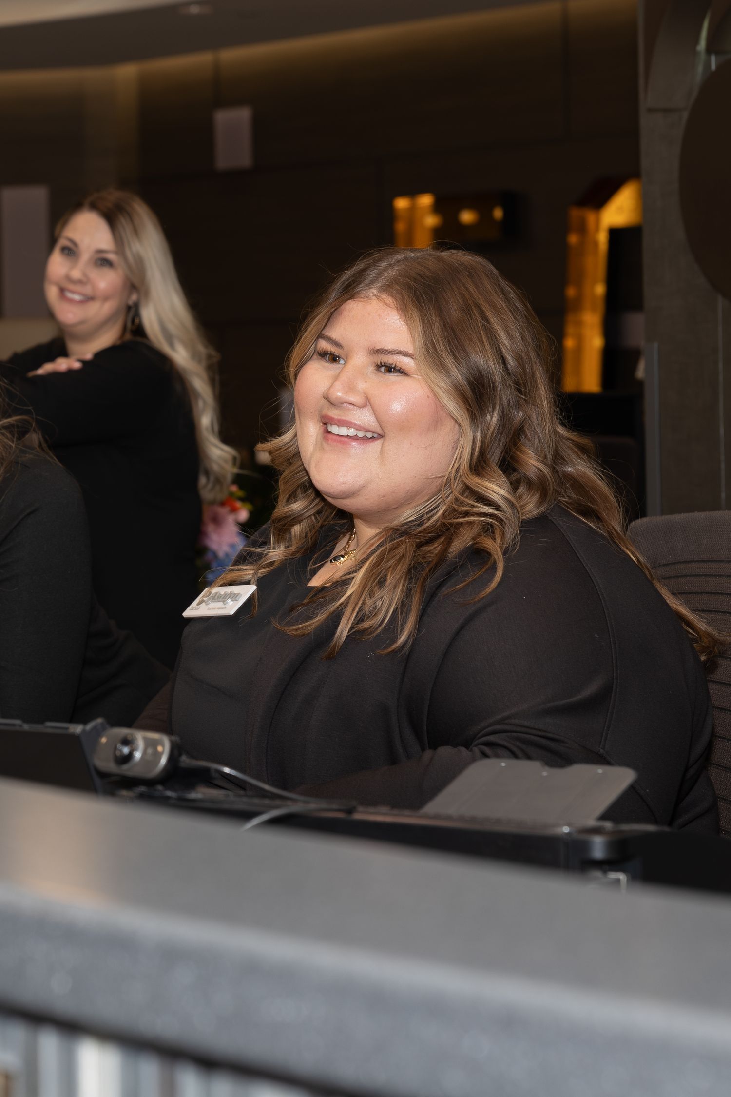 Staff member in black attire smiles warmly while working at front desk reception area.