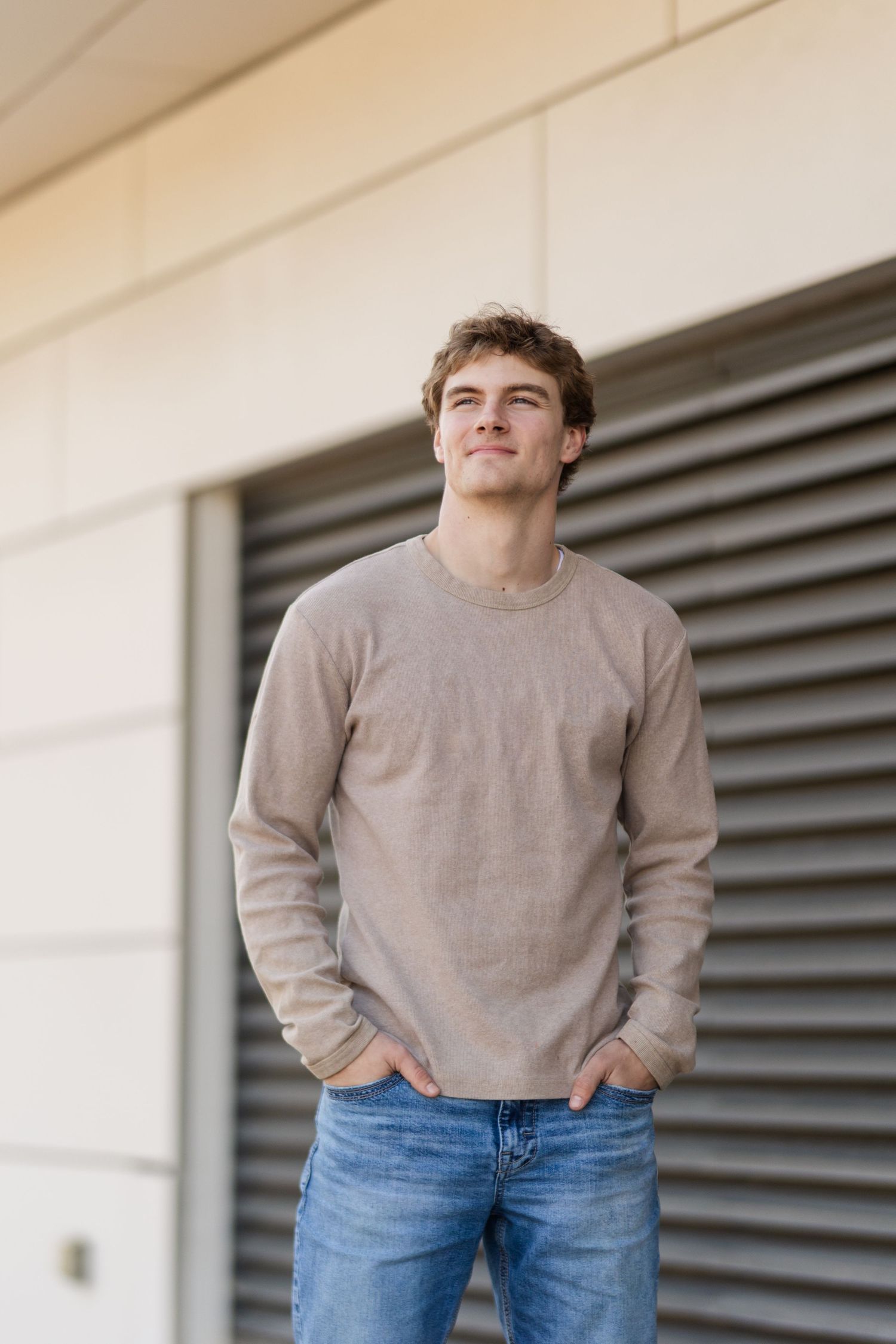 Young person in a beige sweater and jeans stands casually against a wall with modern architectural details.