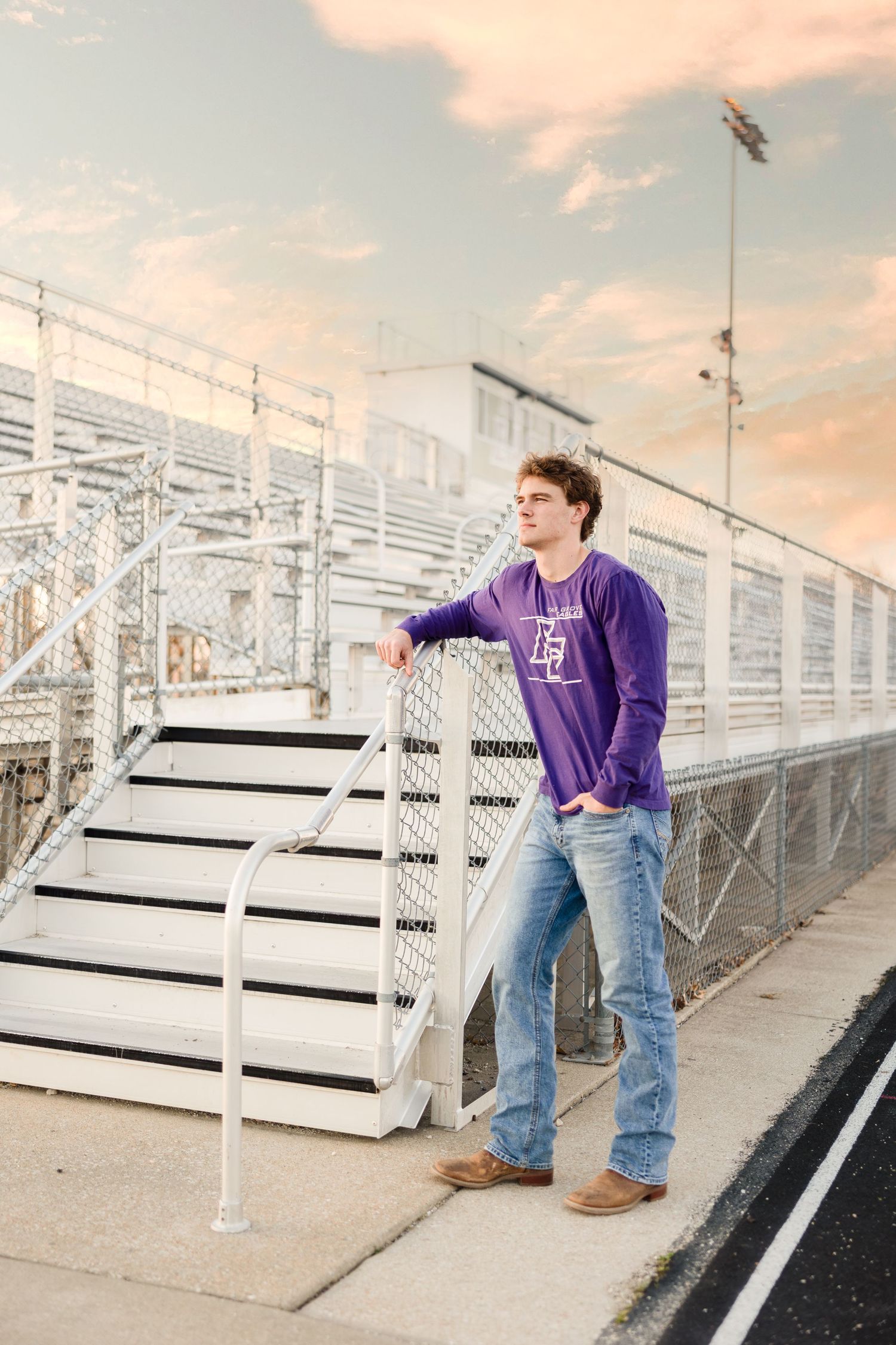 Person in purple sweatshirt and jeans stands by bleachers at sunset against cloudy sky.