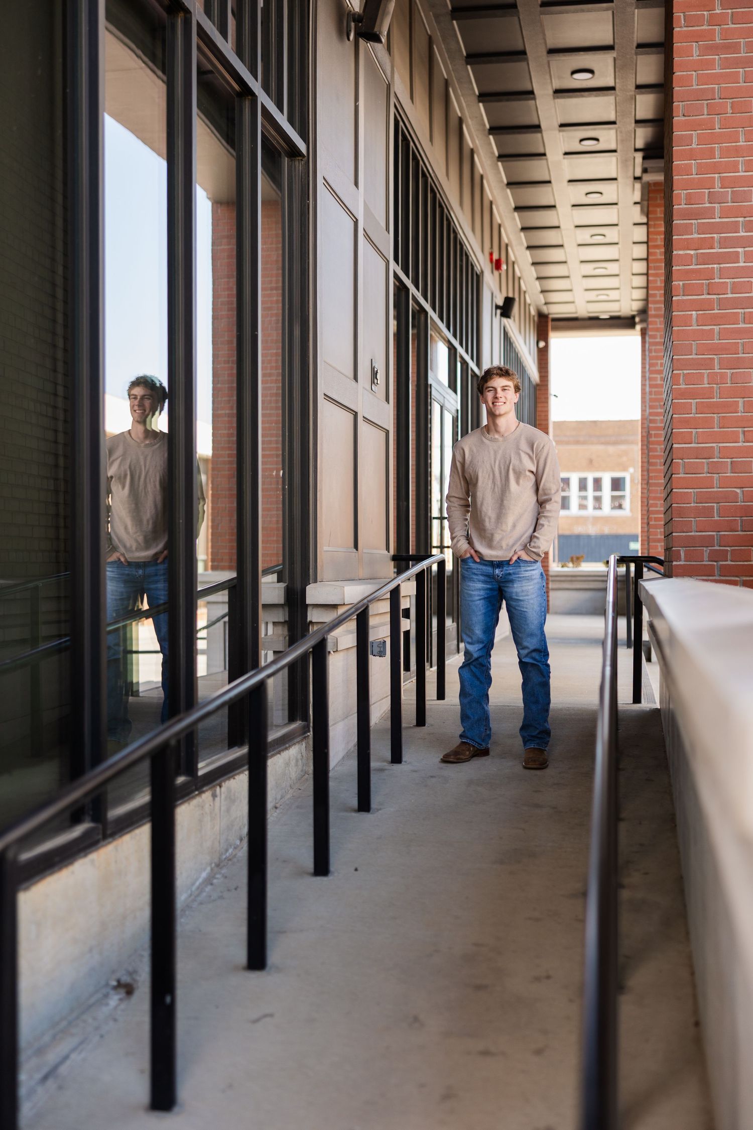 A person in casual attire stands on a covered walkway with brick walls and metal railings.