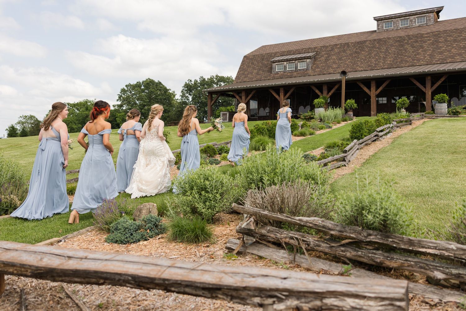 A bridal party in pastel blue dresses walks along a garden path toward a rustic barn venue on a sunny day.