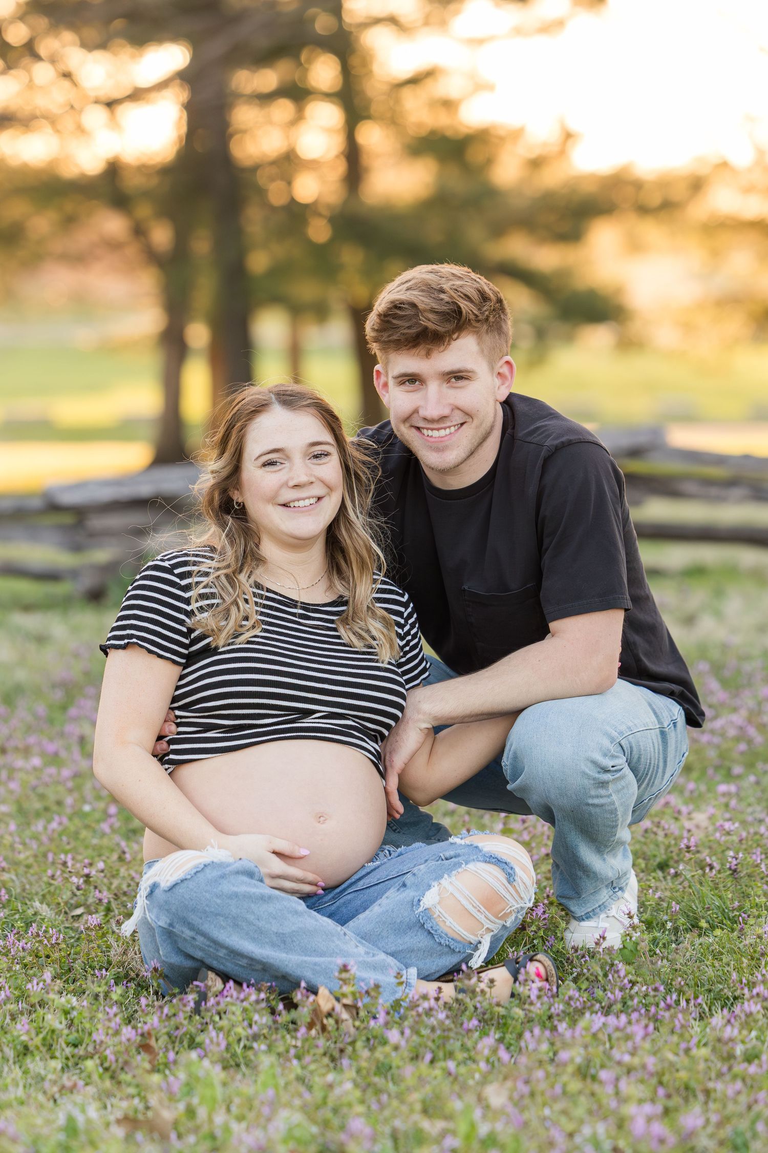 Couple sits together in a field of wildflowers at sunset, expecting mother wearing striped shirt and ripped jeans.