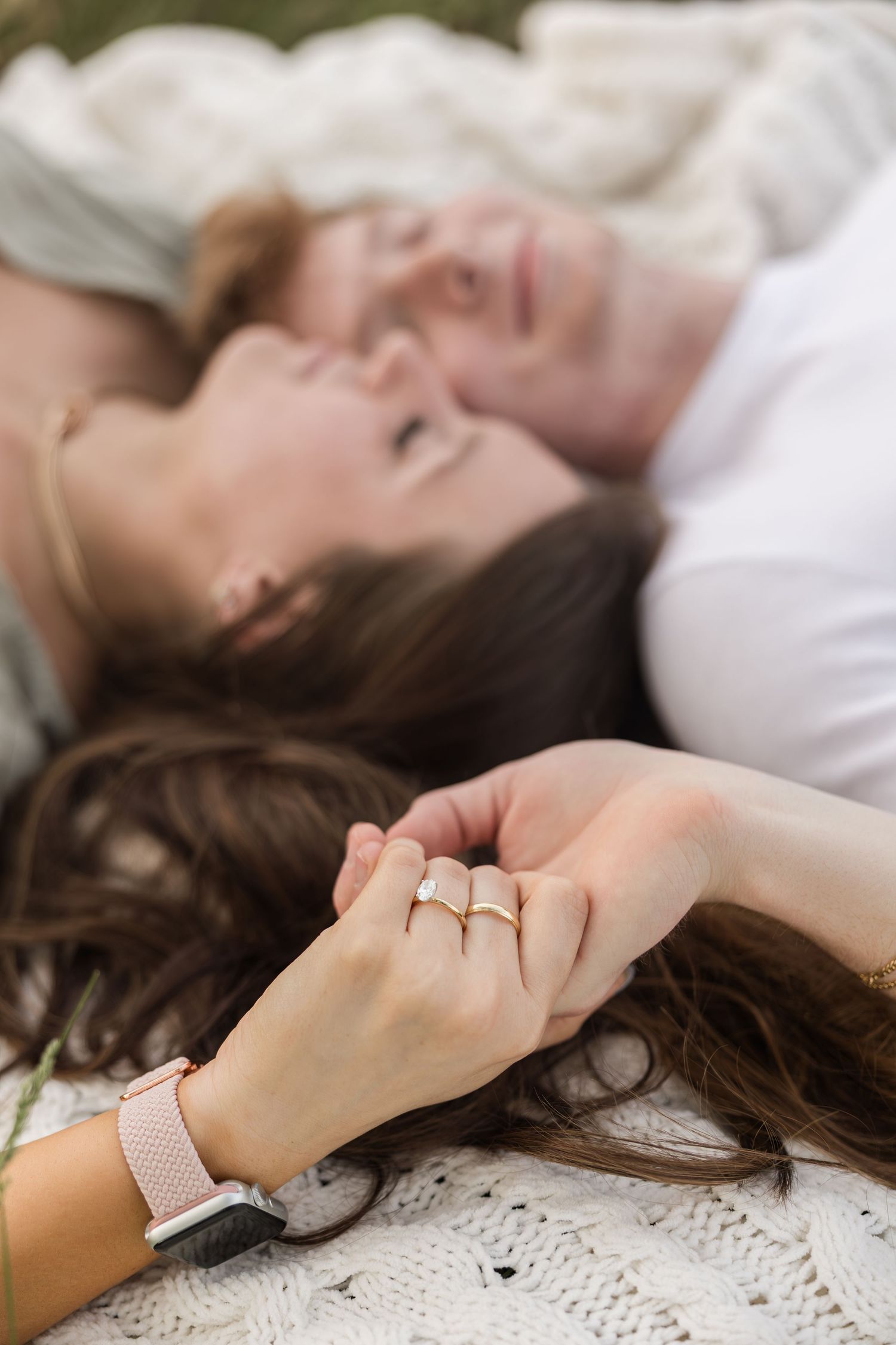 Close up of hands wearing delicate rings holding each other against white bedding in soft light.