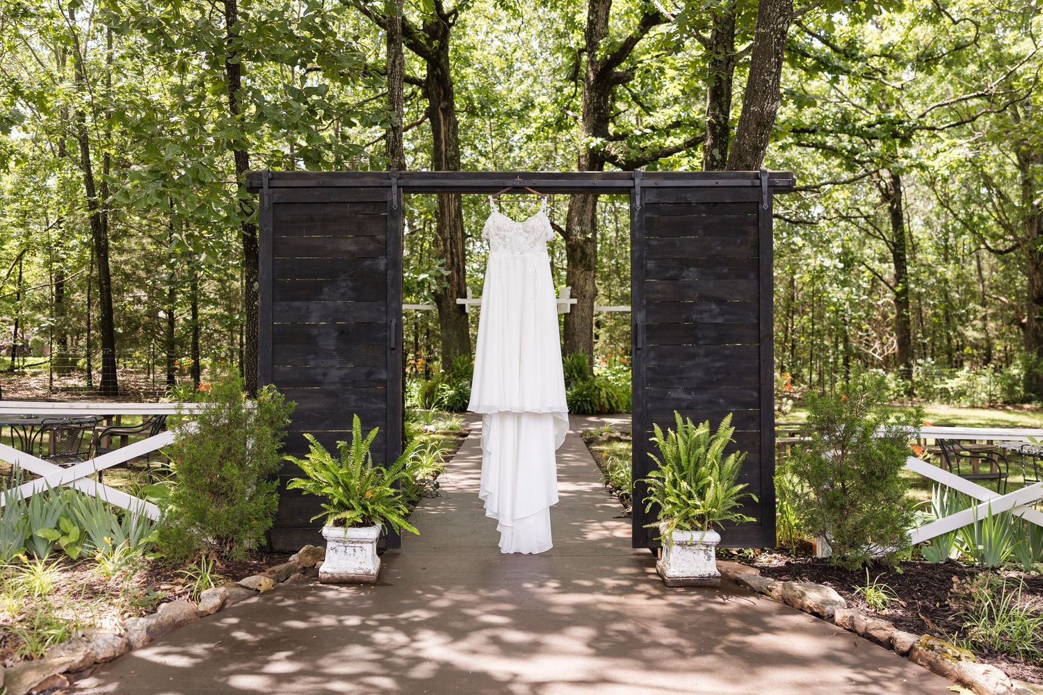 A white wedding dress hangs on display between two black doors in a woodland wedding venue setting with potted plants.