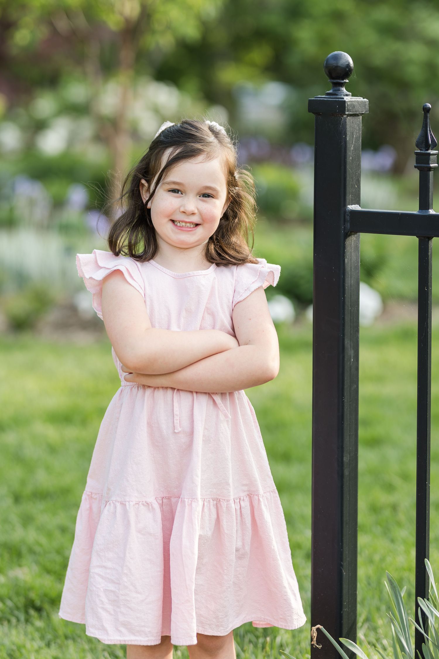 A child in a ruffled pink dress stands next to a black iron garden gate on a spring day.