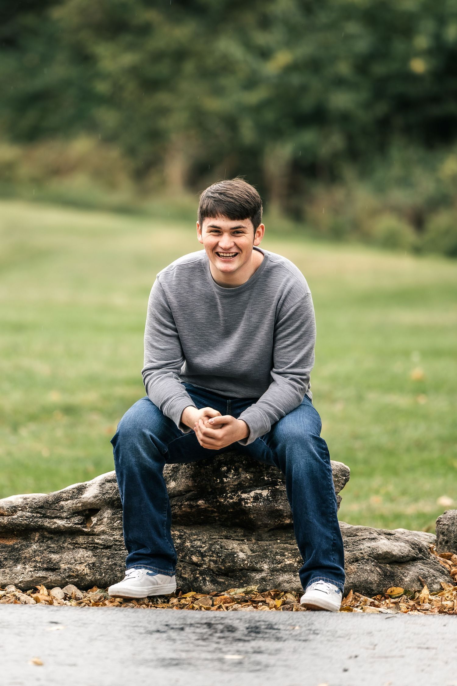Person in gray sweater and jeans sits casually on a fallen log in an autumn outdoor setting.