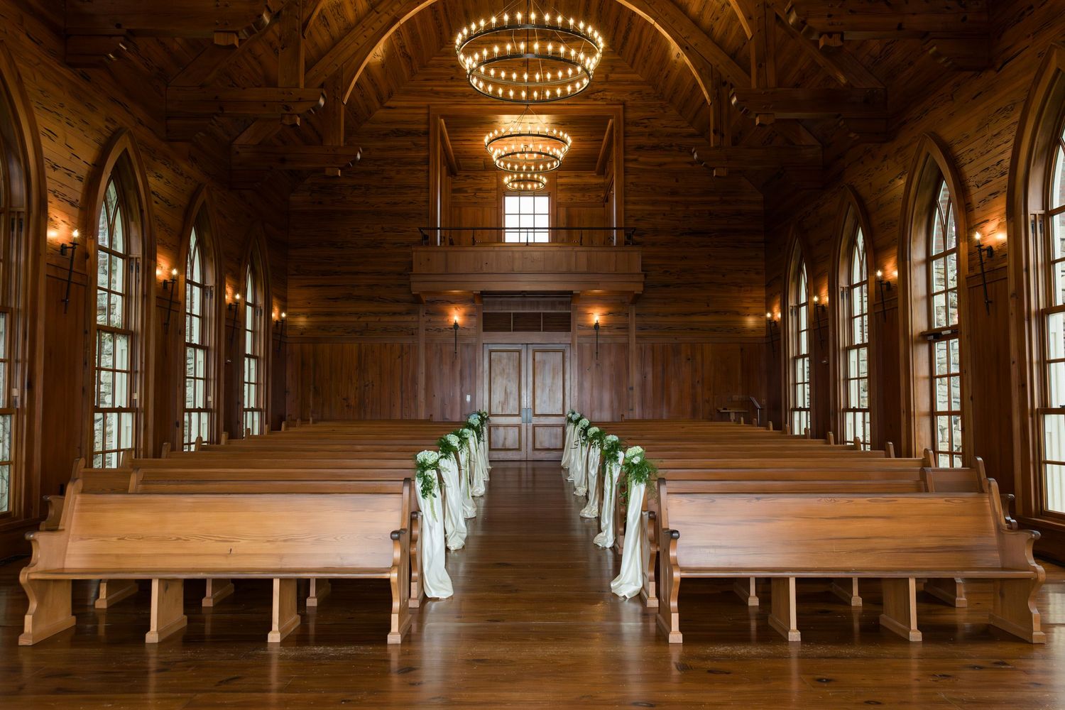 Elegant chandeliers illuminate a grand wooden hall with ornate columns and exposed beams in a historic interior space.