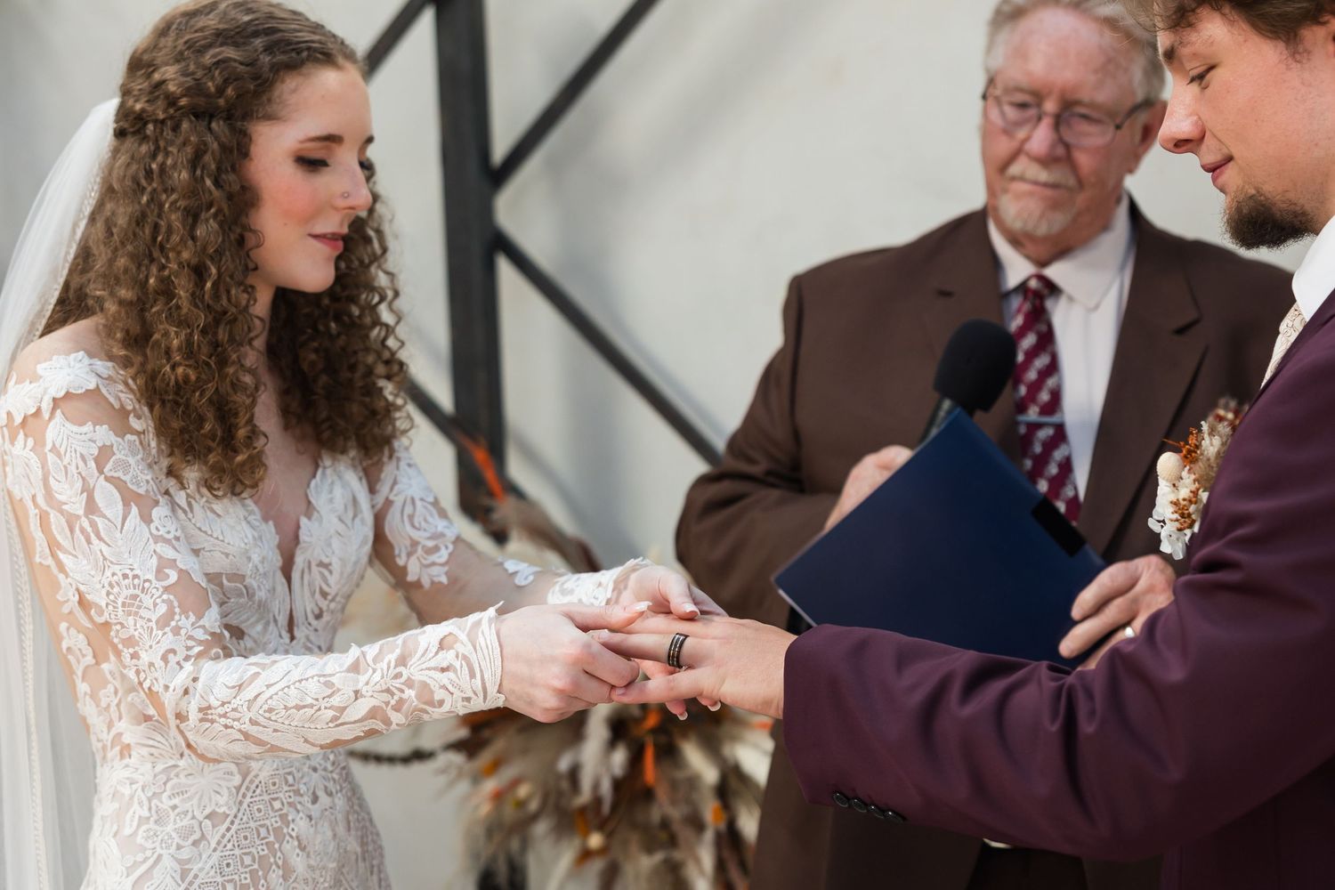 Bride placing wedding ring on groom's finger during ceremony, officiant holding blue book in background.