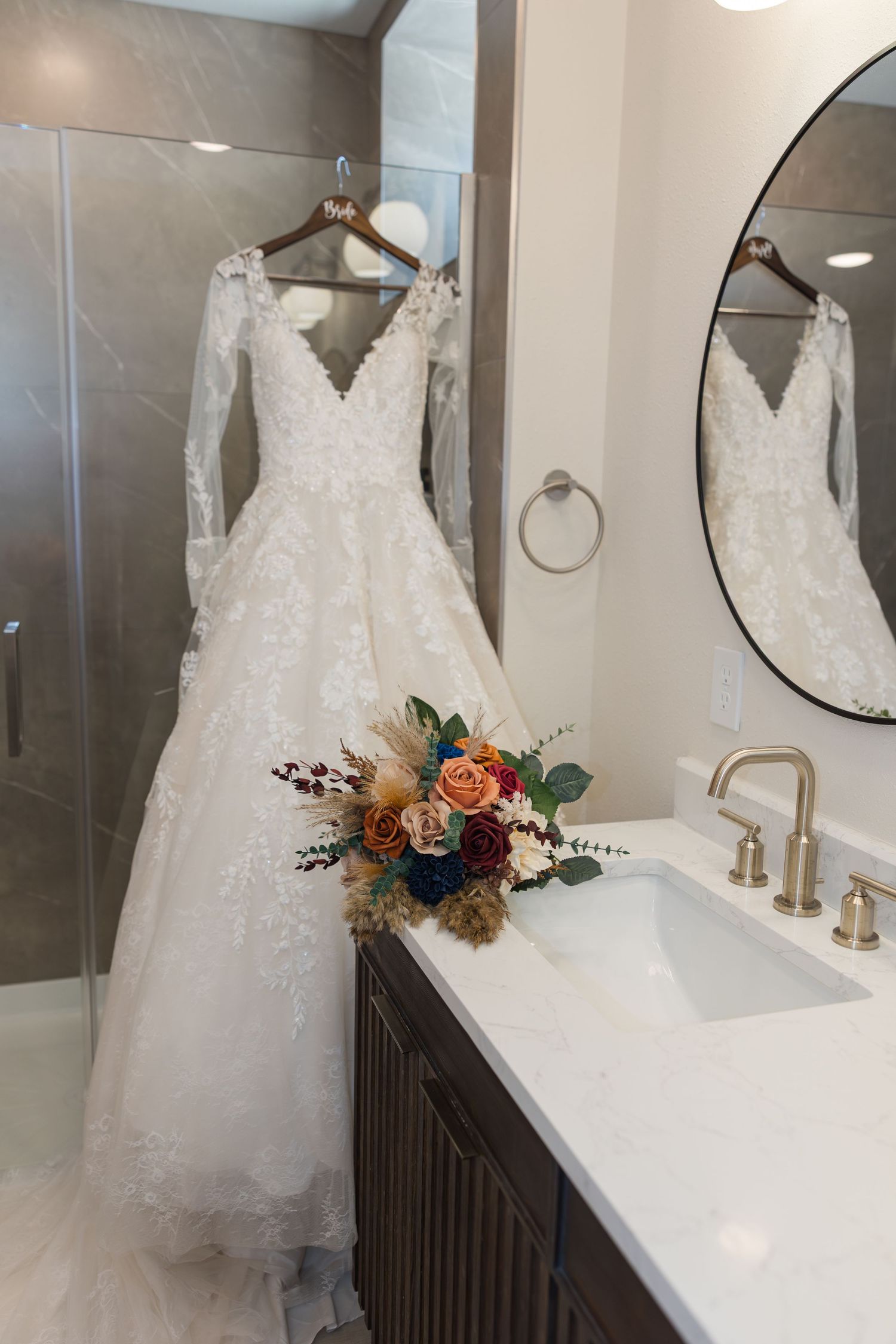 Elegant lace wedding dress with long sleeves hanging in a modern bathroom with a colorful bridal bouquet on the vanity.
