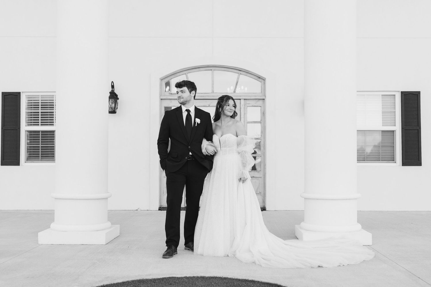 Black and white photo of a bride and groom standing elegantly before white columns at a classic wedding venue.