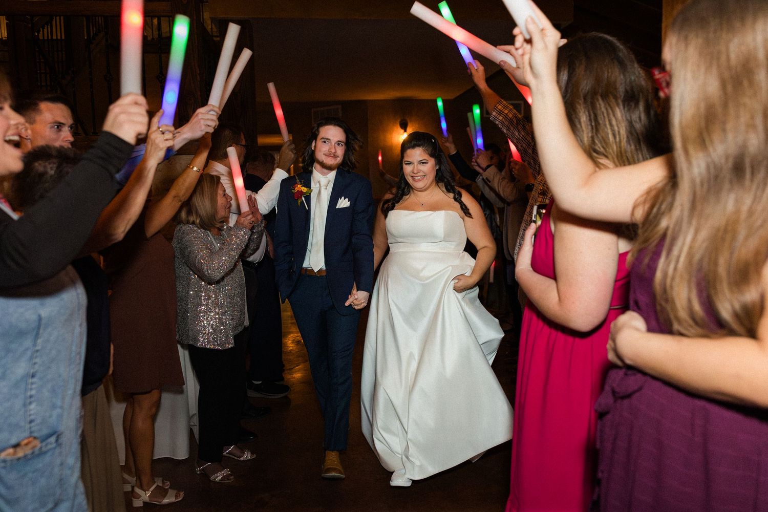 Bride and groom walk through a glowing foam stick tunnel surrounded by cheering wedding guests.