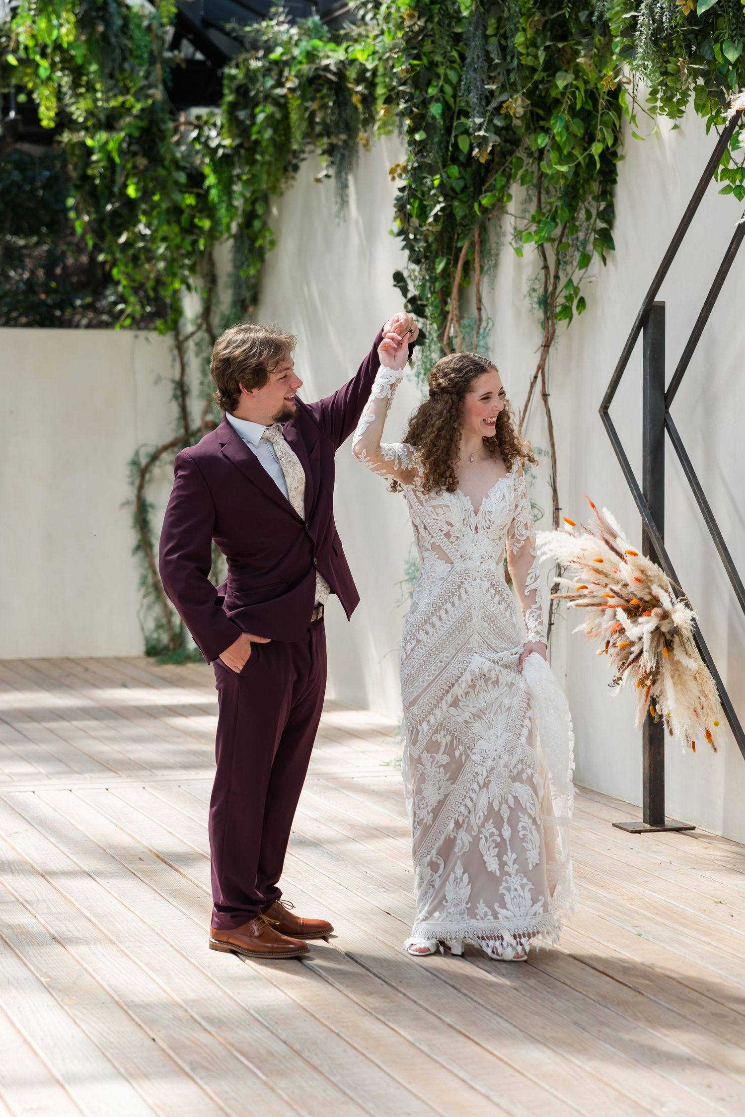 Bride in lace gown twirls as groom in burgundy suit holds her hand on a wooden deck with greenery.