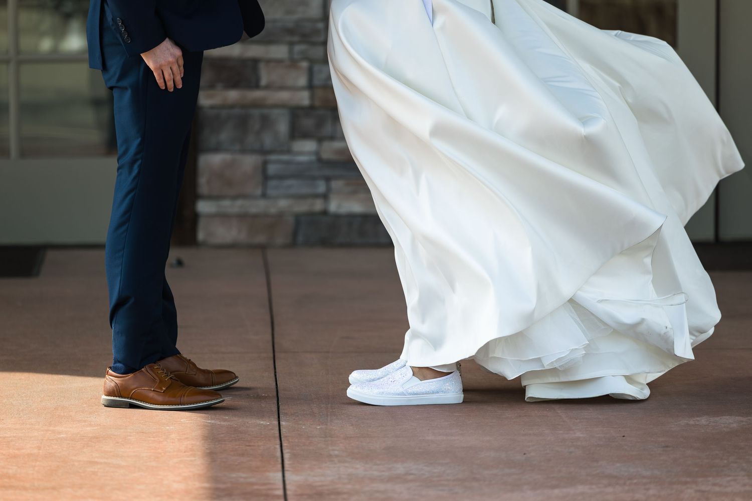 A close-up view of formal dress shoes and a white wedding gown hem on a tiled floor.