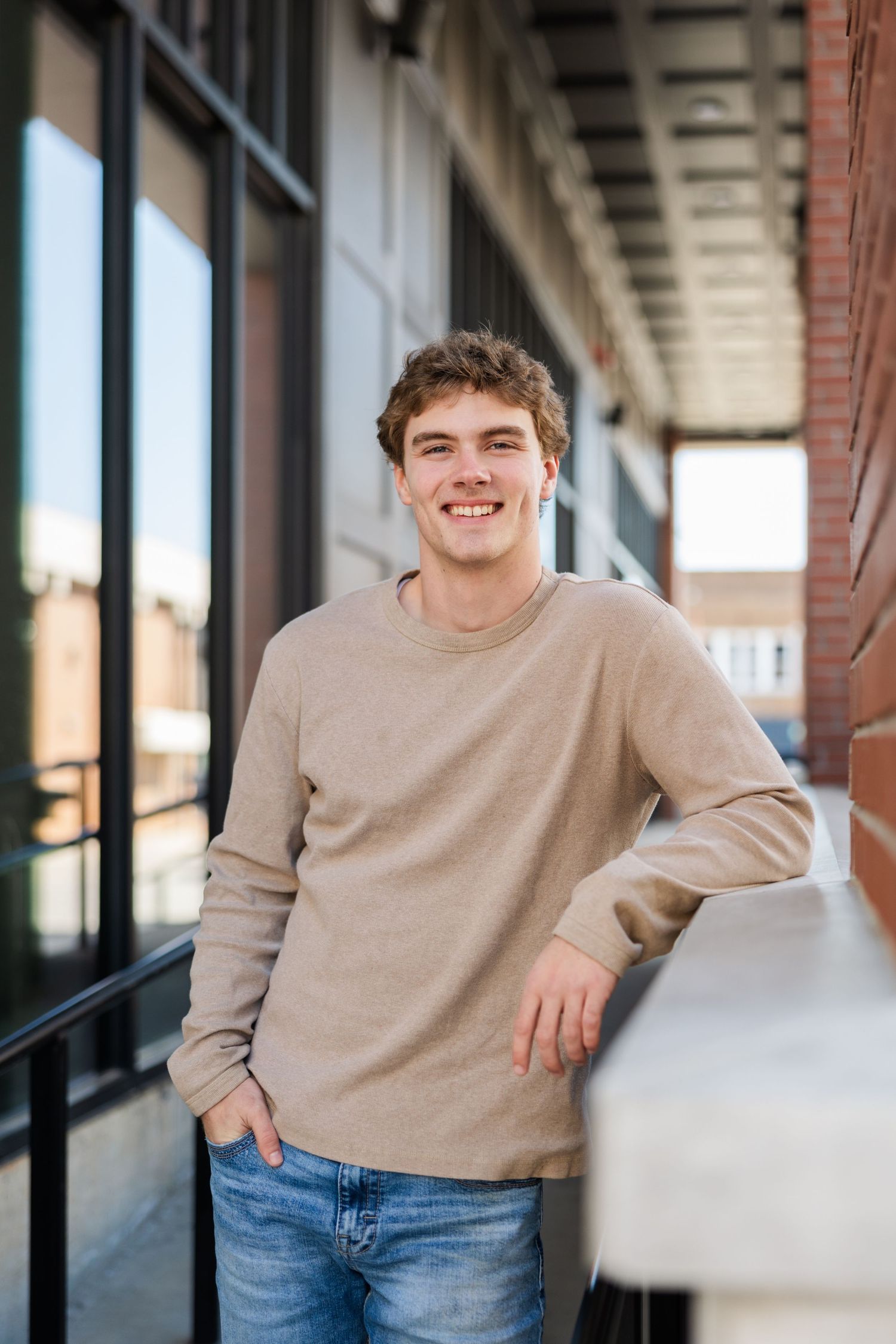 Young person in beige sweater and jeans leaning casually against wall in urban setting with bright smile.