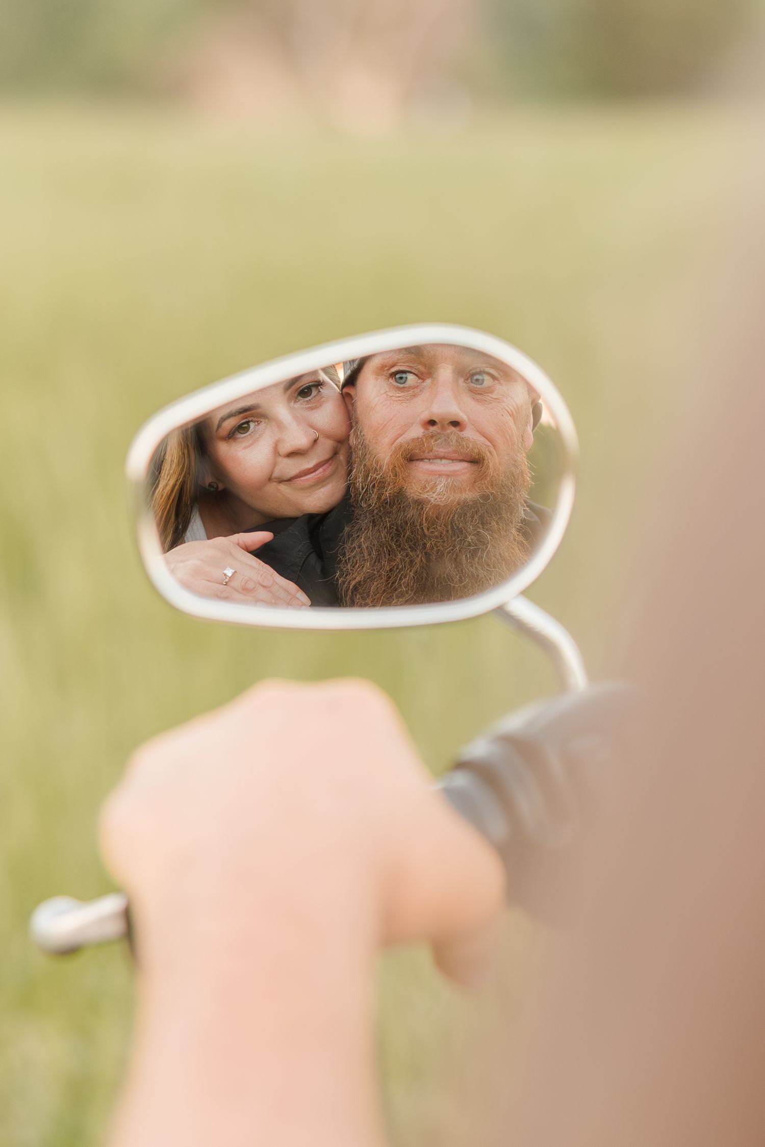 A creative reflection in a motorcycle side mirror shows a couple smiling against a soft, blurred outdoor background.