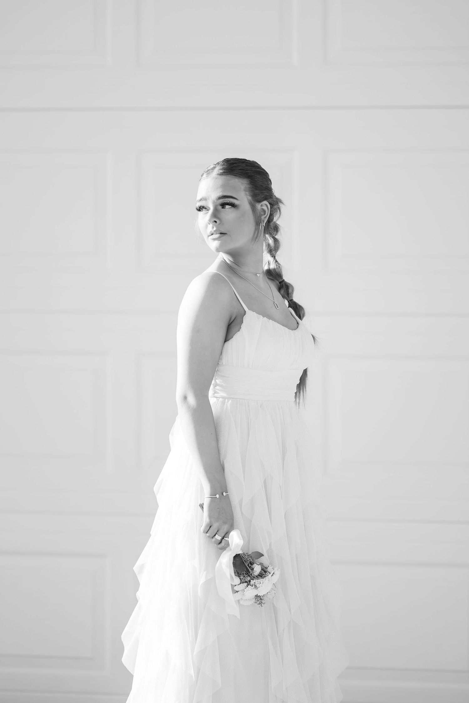 A black and white portrait of a bride in a flowing white dress with a braided hairstyle against a light background.