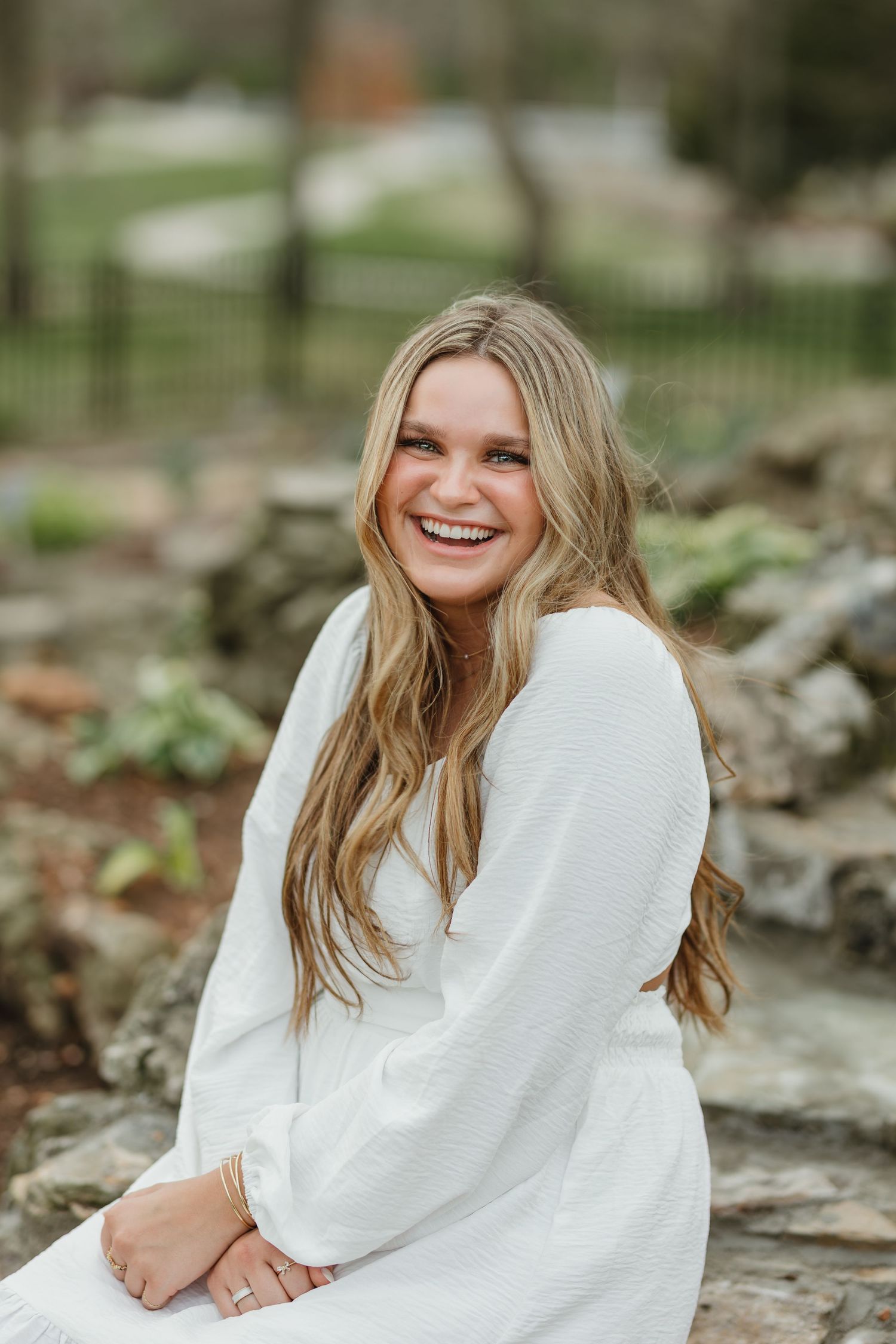 A person wearing a white dress smiles while sitting on rocks in an outdoor setting.
