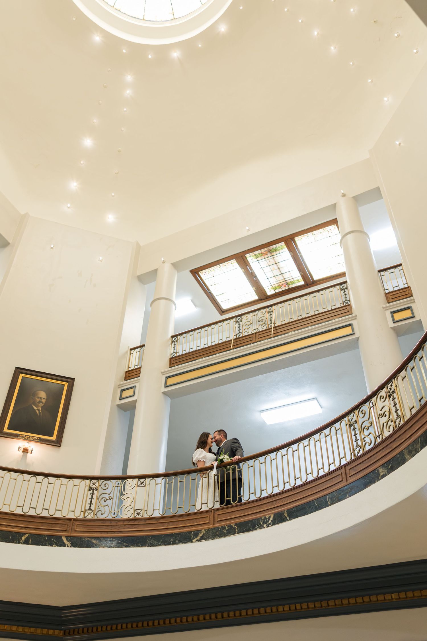 A couple stands on an elegant curved balcony inside a historic building with a domed ceiling and white columns.