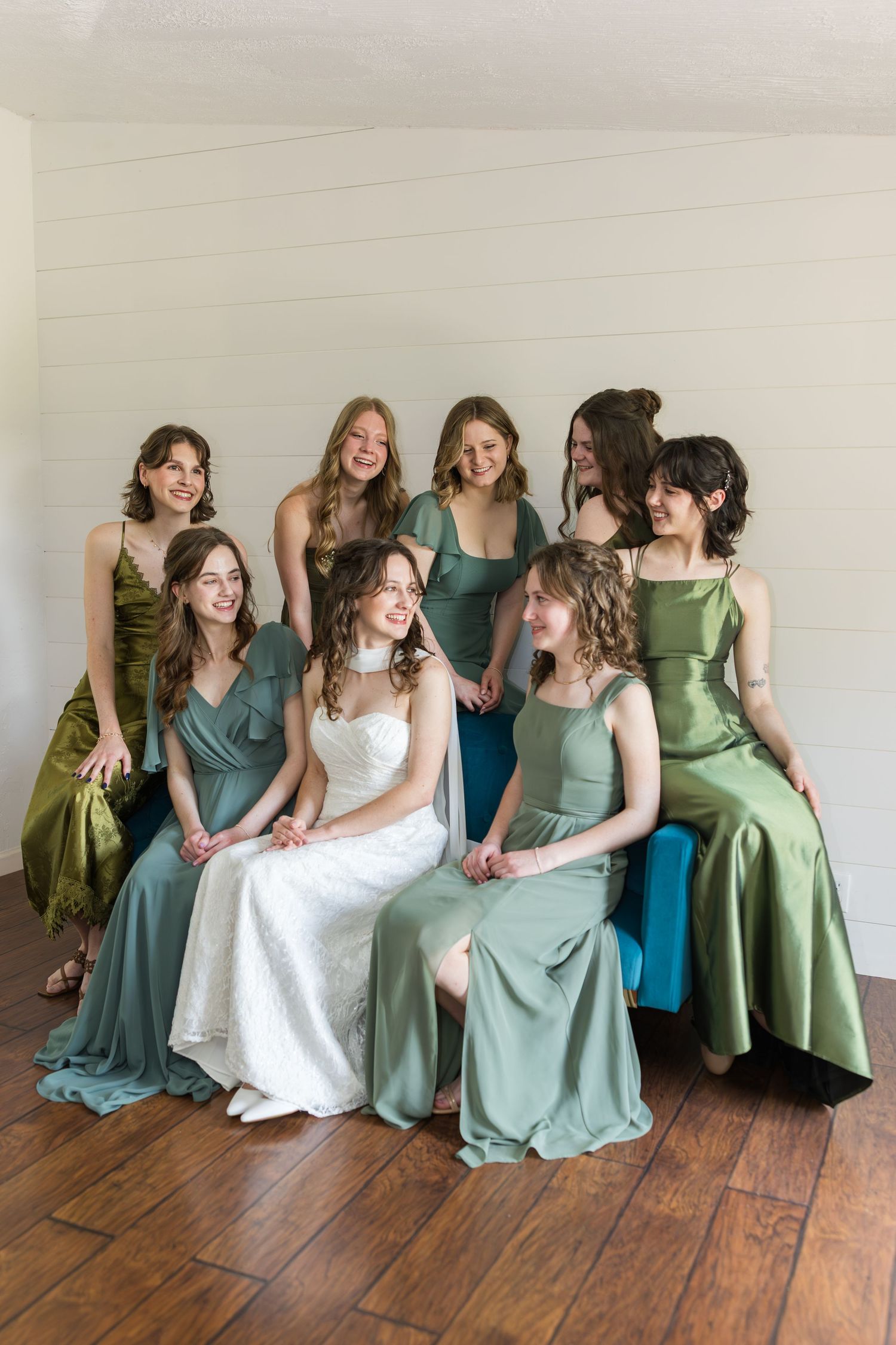 Bride in white lace gown poses with seven bridesmaids in mismatched green satin dresses, smiling together indoors.