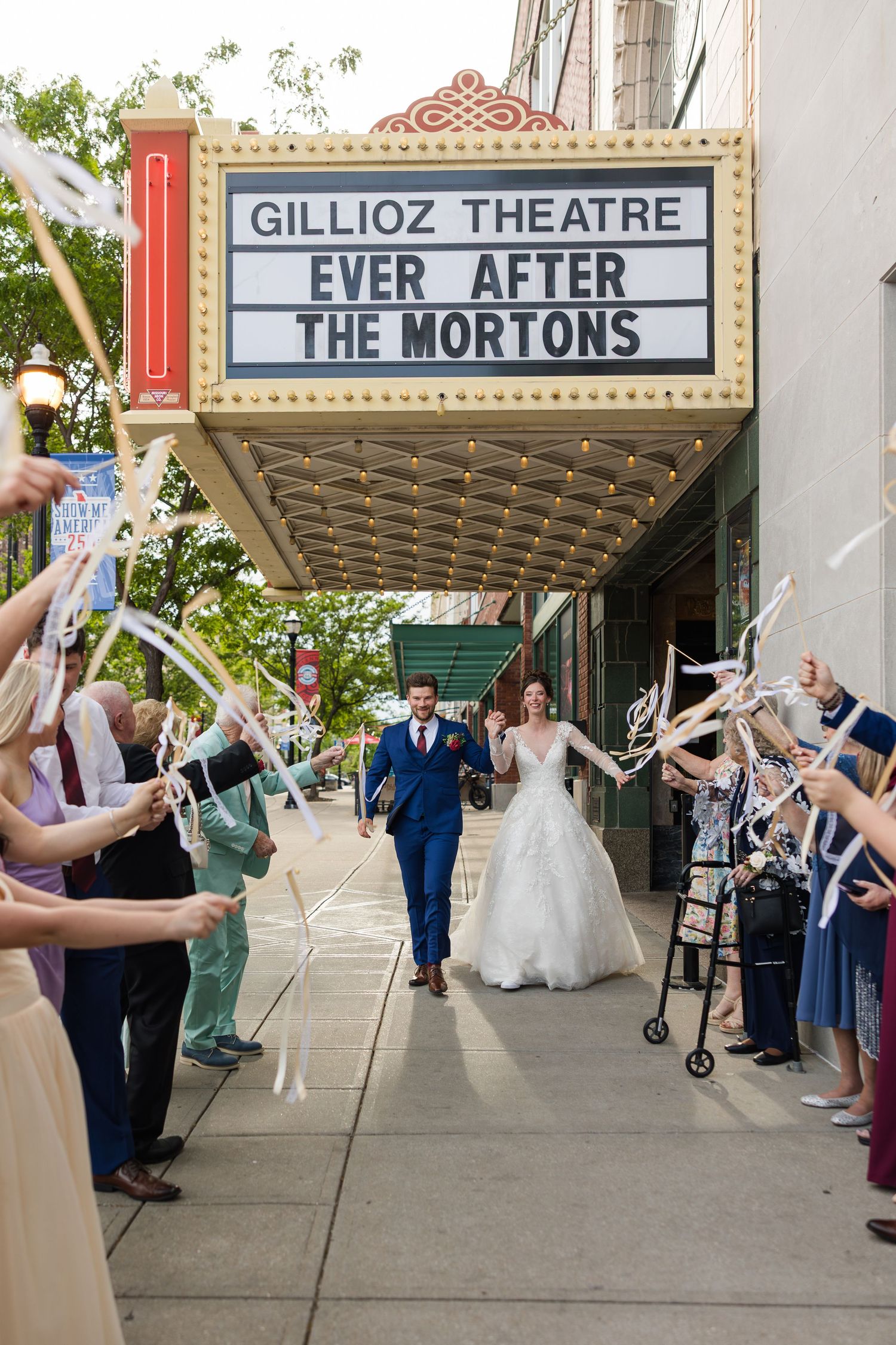Newlyweds exit Gillioz Theatre under marquee reading Ever After The Mortons amid ribbon wands.