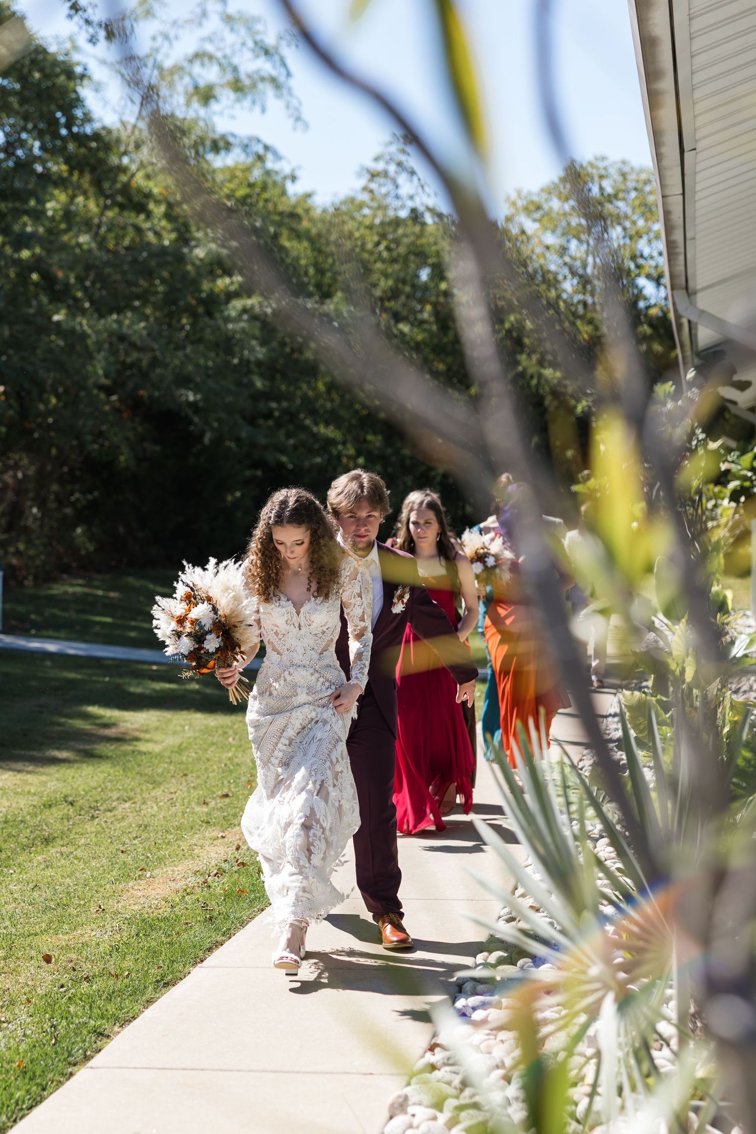 Bride and groom walk down outdoor path with bridesmaid in red dress carrying bouquets on sunny wedding day.