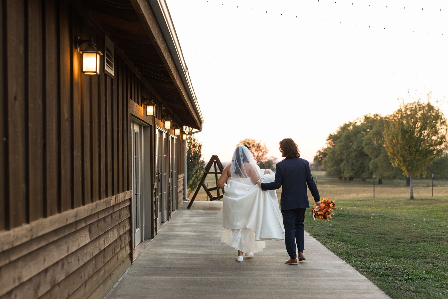 Bride and groom walking hand in hand along a rustic barn venue pathway at golden hour sunset.