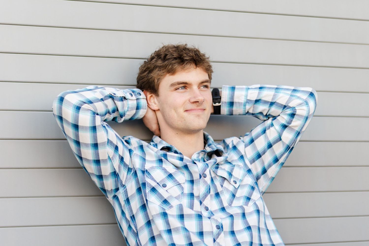 Person in blue plaid shirt leaning back with hands behind head against gray wall.