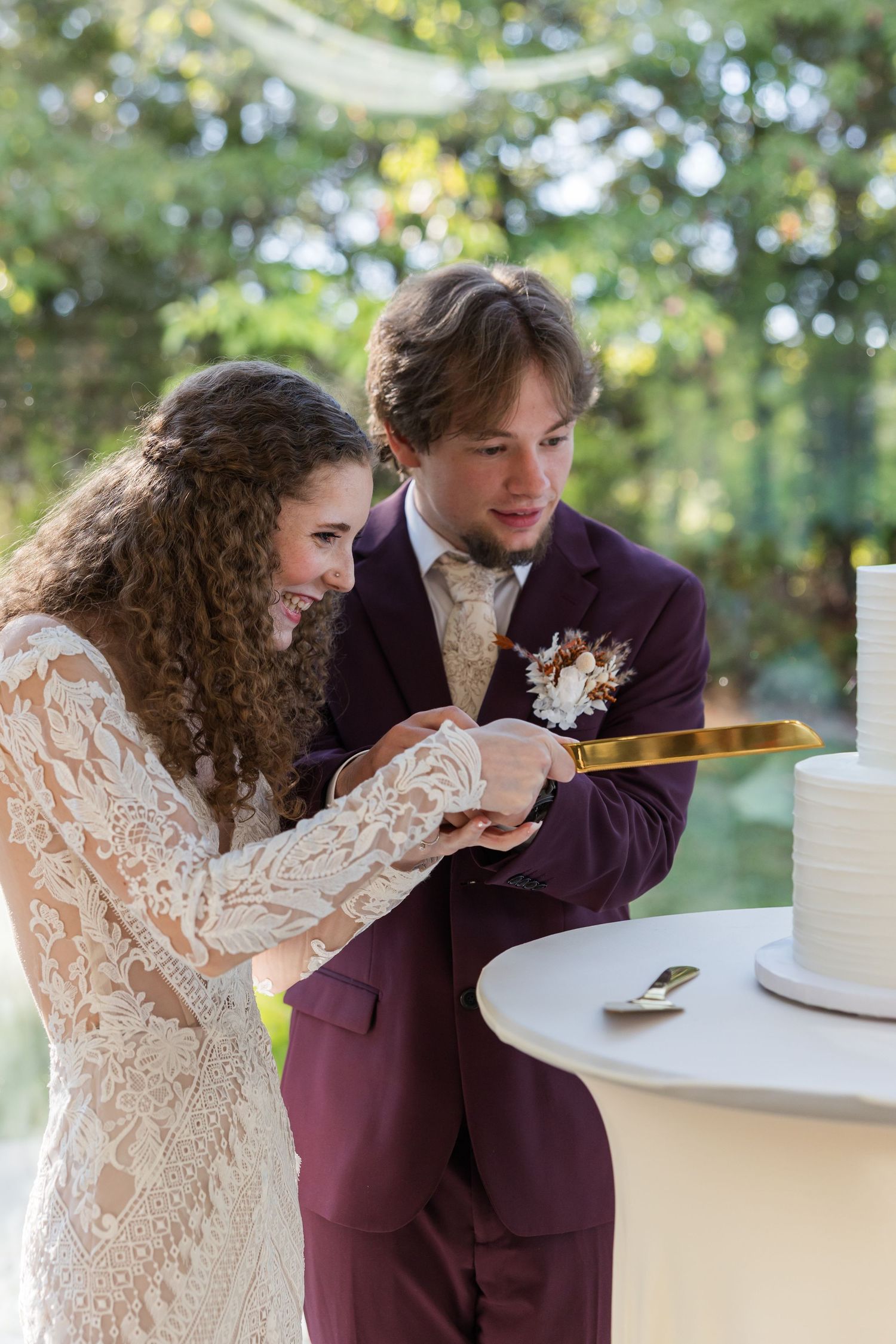 A couple in wedding attire share a sweet moment while cutting their white wedding cake together at an outdoor reception.